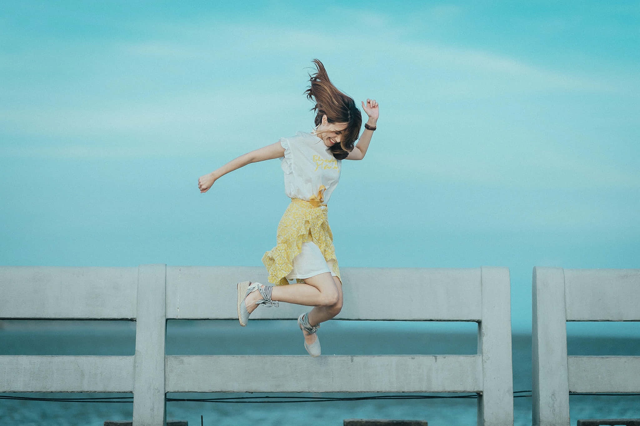 happiness 桌布 壁紙 — 一個快樂的女孩在廣闊的藍綠色天空和海洋背景下，跳過混凝土欄杆，騰空而起。