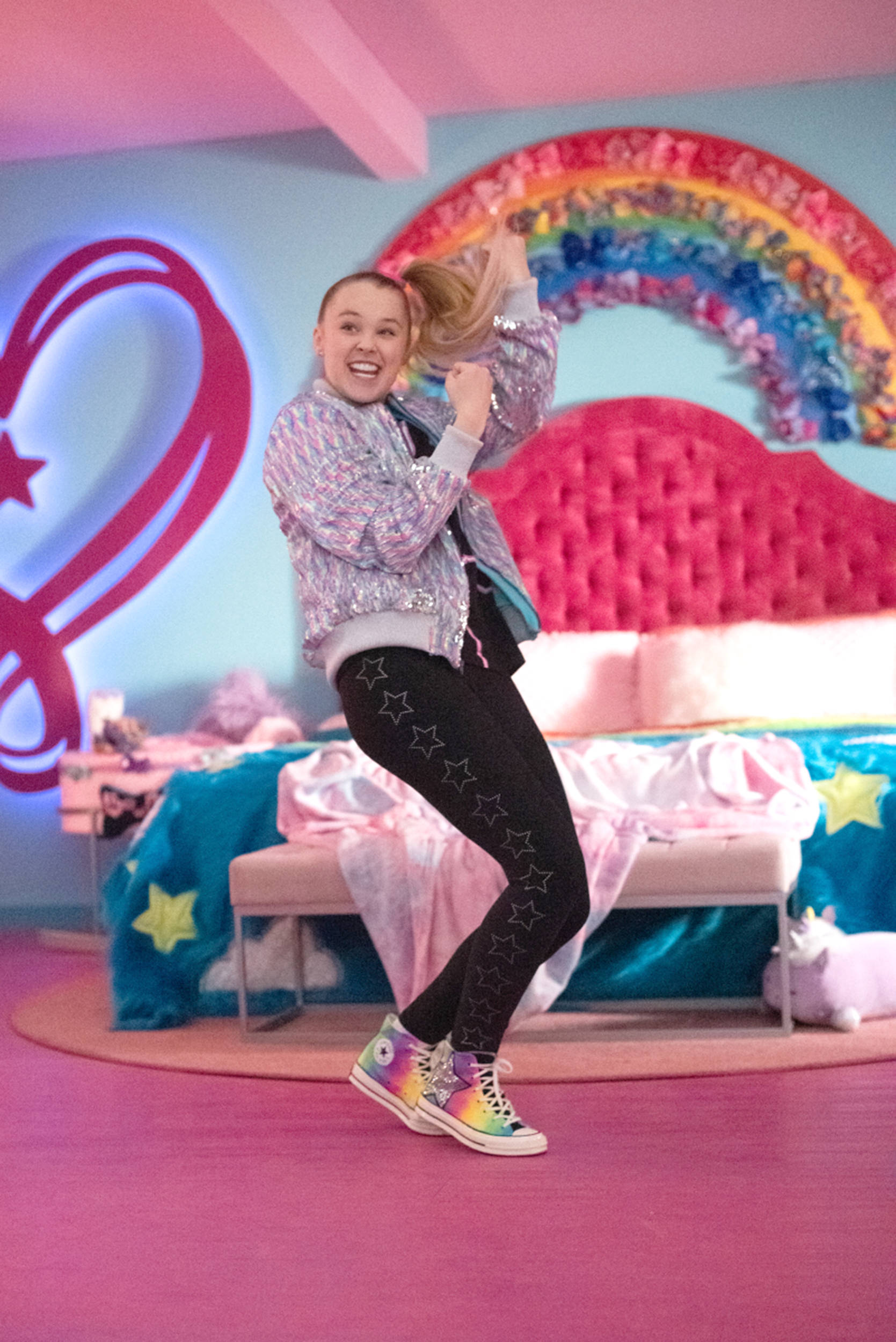 JoJo Siwa 桌布 壁紙 — 一個快樂的女孩穿著閃亮外套和星星緊身褲，在充滿活力的粉紅色房間裡，配著彩虹床頭板，充滿活力地跳舞。