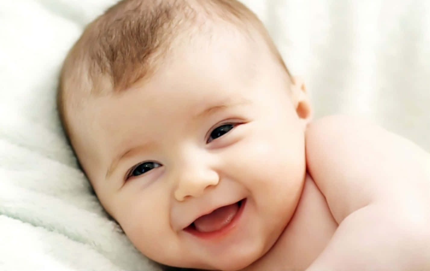 Baby 桌布 壁紙 — 一個快樂的嬰兒，深色眼睛和柔軟的棕色頭髮，躺在柔軟的白色毯子上，露出燦爛的笑容。