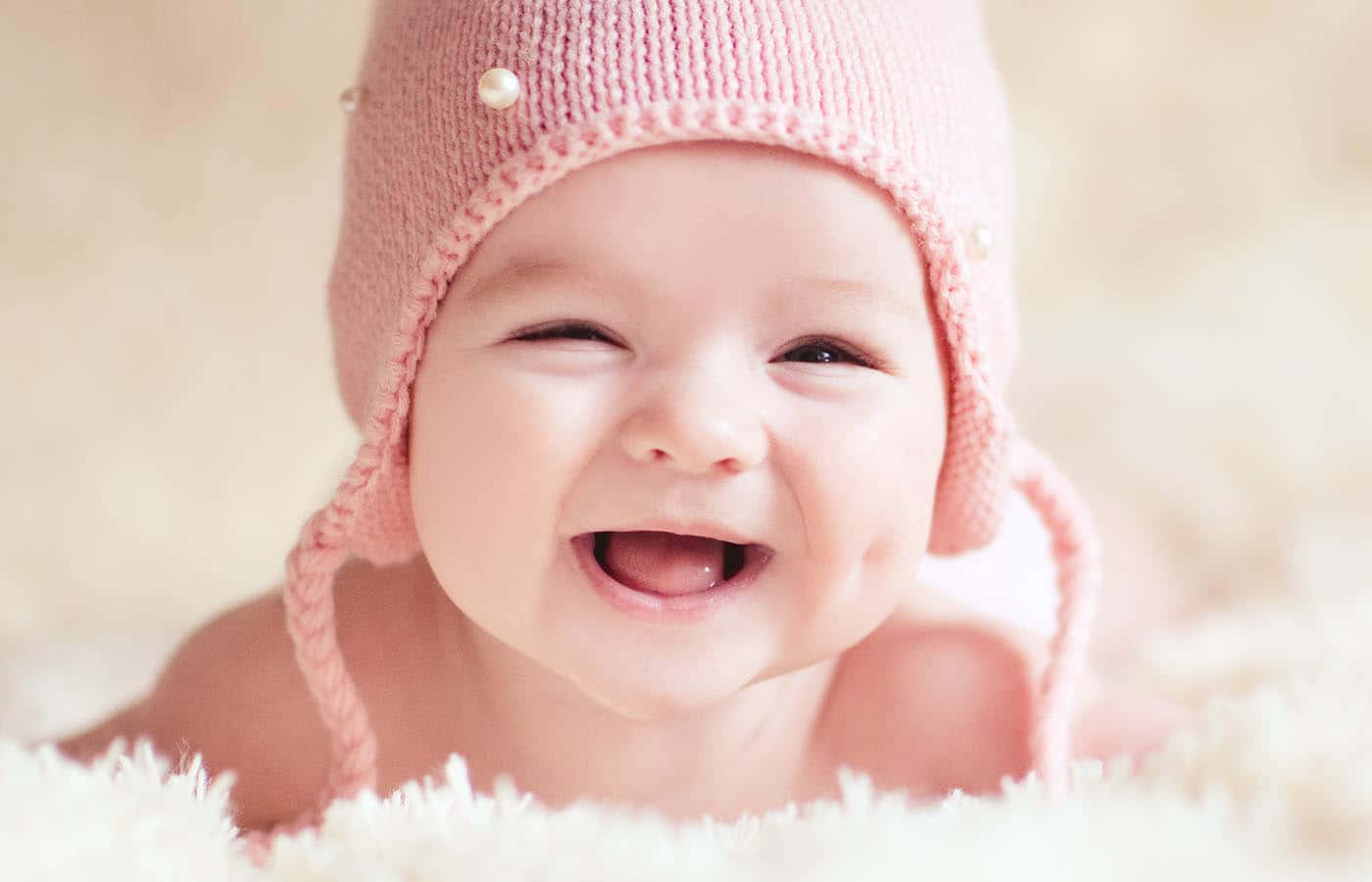 Contentment 桌布 壁紙 — 一個快樂的嬰兒在柔軟的白色表面上戴著帶有珍珠裝飾的柔軟粉紅色針織帽，開懷大笑。