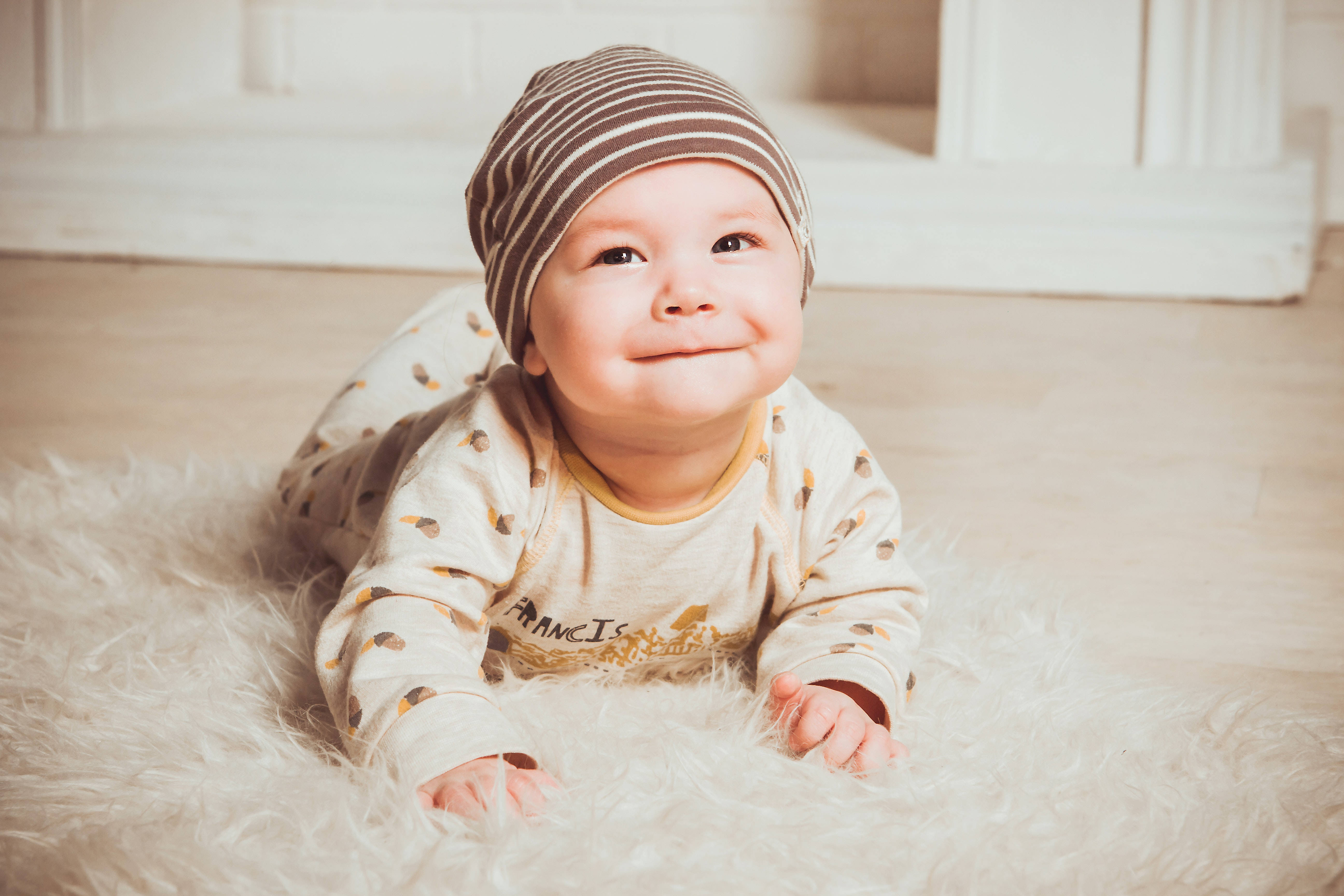 Smile 桌布 壁紙 — 一個快樂的嬰兒戴著條紋帽，躺在室內柔軟的白色地毯上微笑。