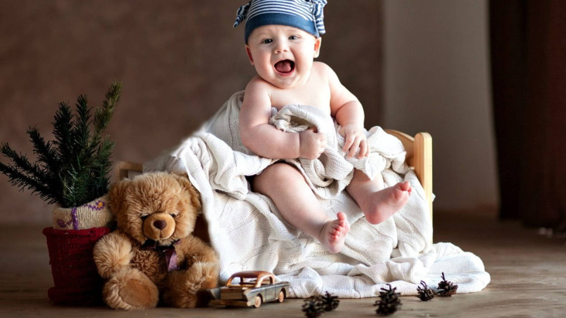 Baby 桌布 壁紙 — 一個快樂的嬰兒戴著條紋帽子，坐在木椅上，裹著白色毯子，周圍有泰迪熊、松果和玩具車。