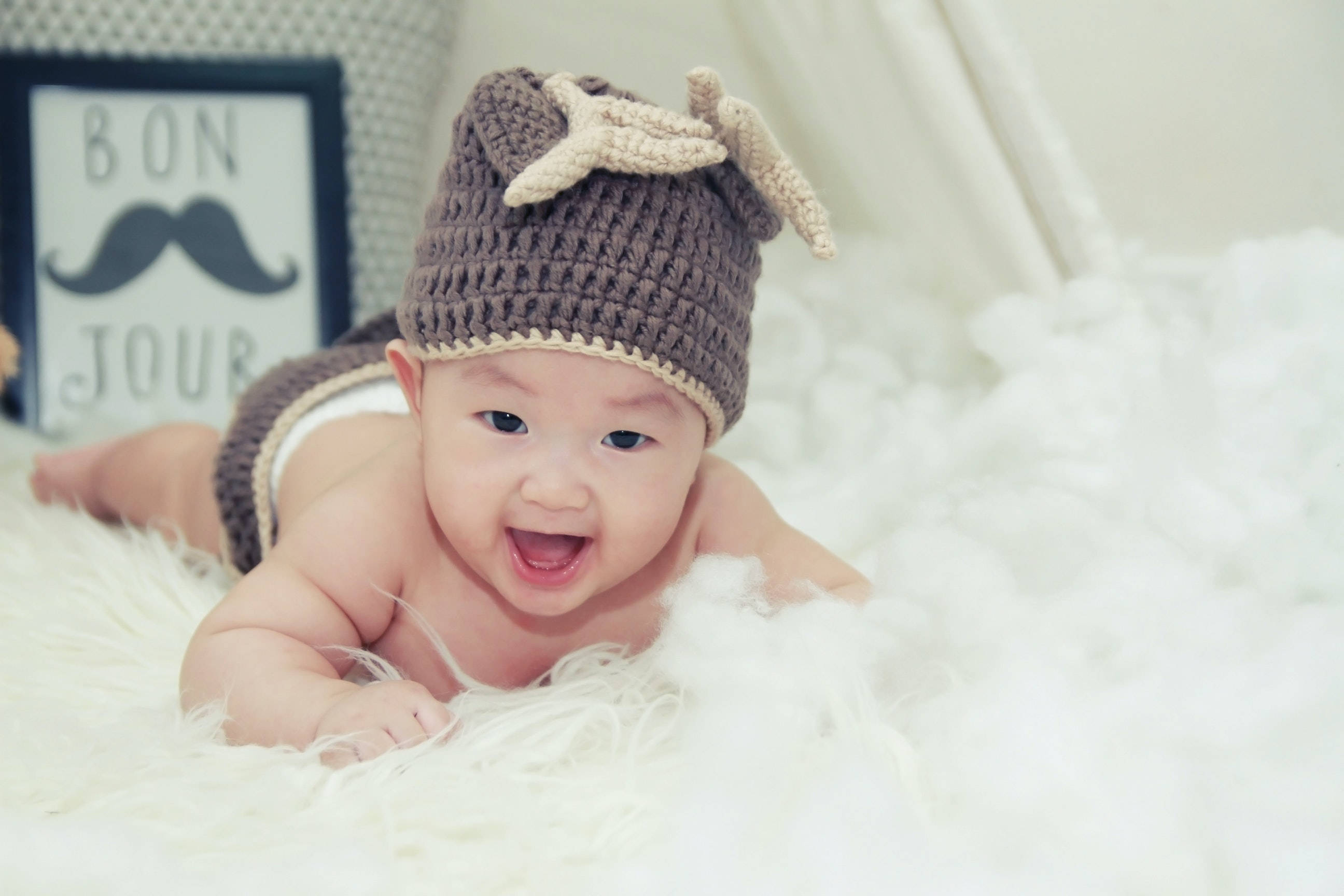 Very Cute Baby 桌布 壁紙 — 一個快樂的嬰兒戴著棕色鉤針編織帽，上面有海星和鹿角細節，躺在柔軟的白色地毯上。