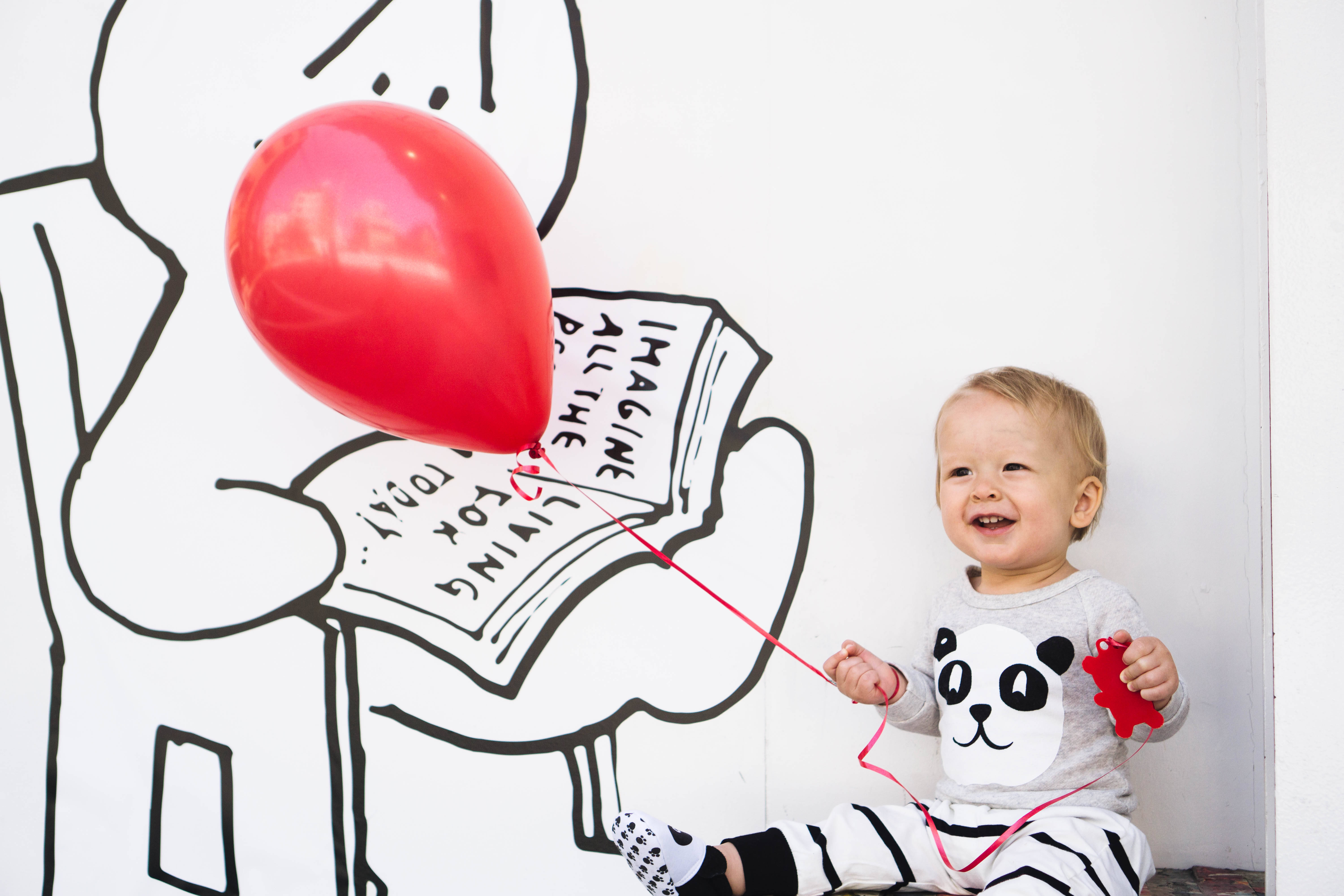 Baby 桌布 壁紙 — 一個快樂的嬰兒穿著熊貓睡衣，笑著拿著一個繫在巨大插圖書壁畫上的紅色氣球。