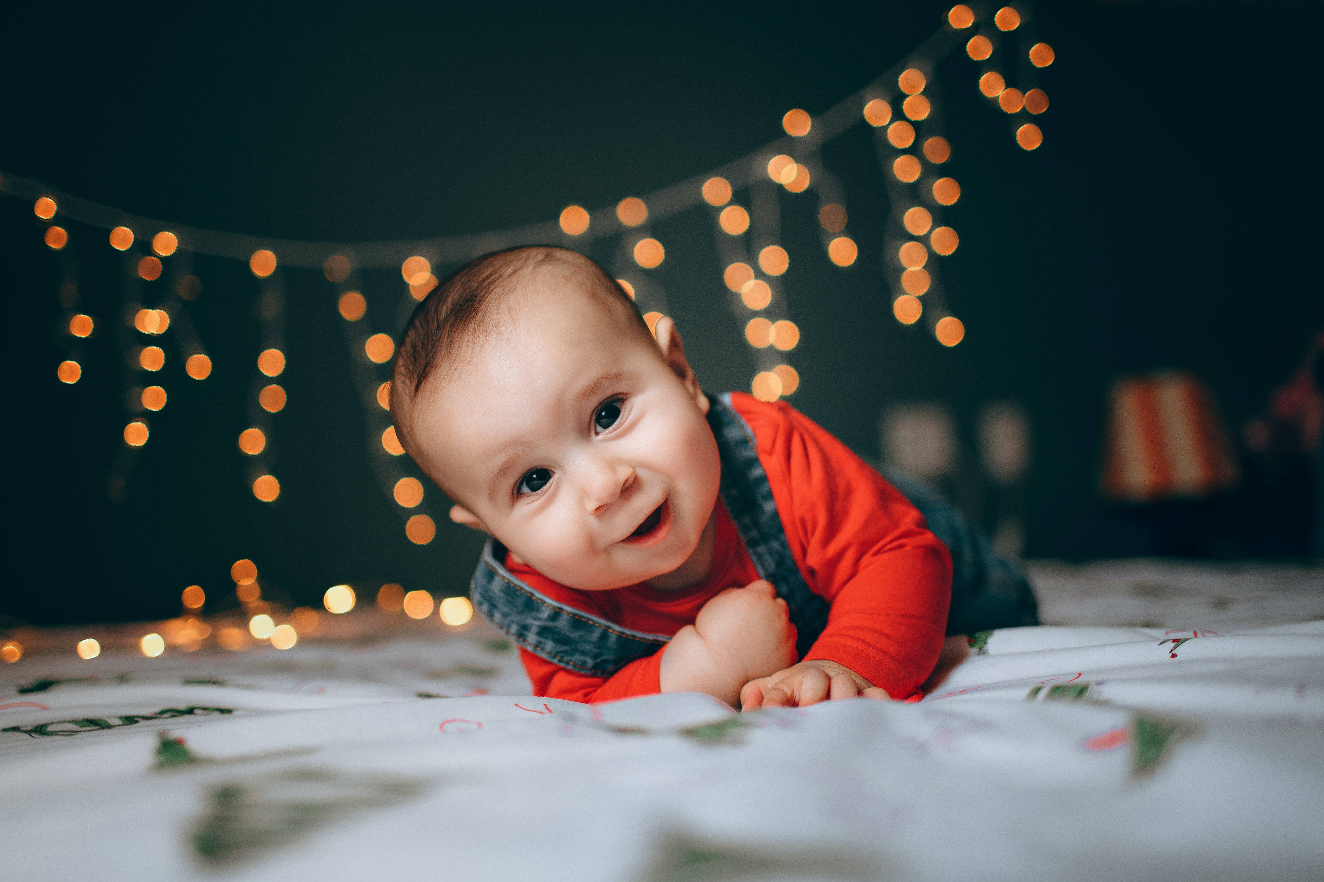 Very Cute Baby 桌布 壁紙 — 一個快樂的嬰兒穿著紅色襯衫和牛仔布工裝褲，在溫暖的串燈下爬行在圖案毯子上微笑。