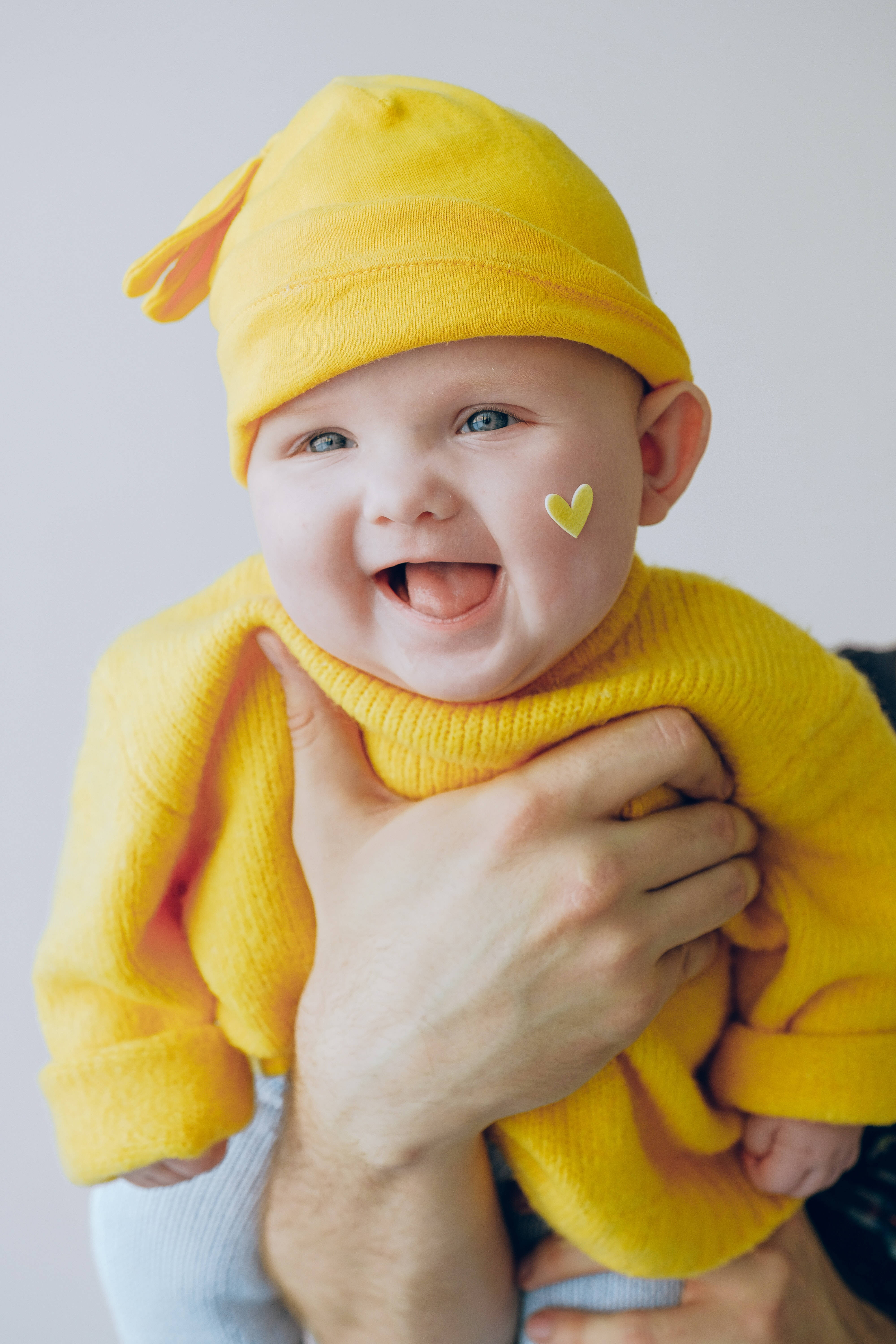 Baby Boy 桌布 壁紙 — 一個快樂的嬰兒穿著鮮黃色衣服和帽子，被成年人的手抱著，在純色背景上露出燦爛的笑容。