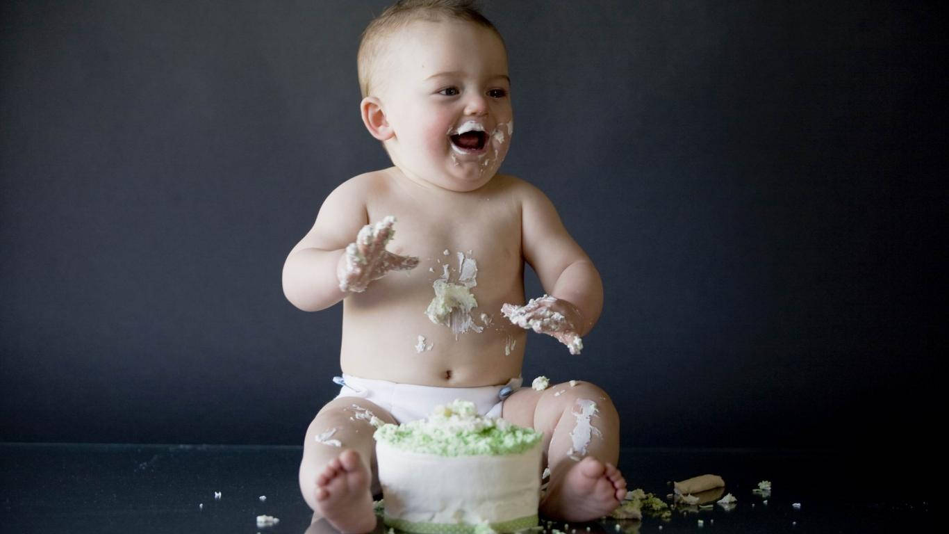 Funny Baby 桌布 壁紙 — 一個快樂的嬰兒被白色糖霜覆蓋，在深色背景前開心地坐著，旁邊有一個小蛋糕。