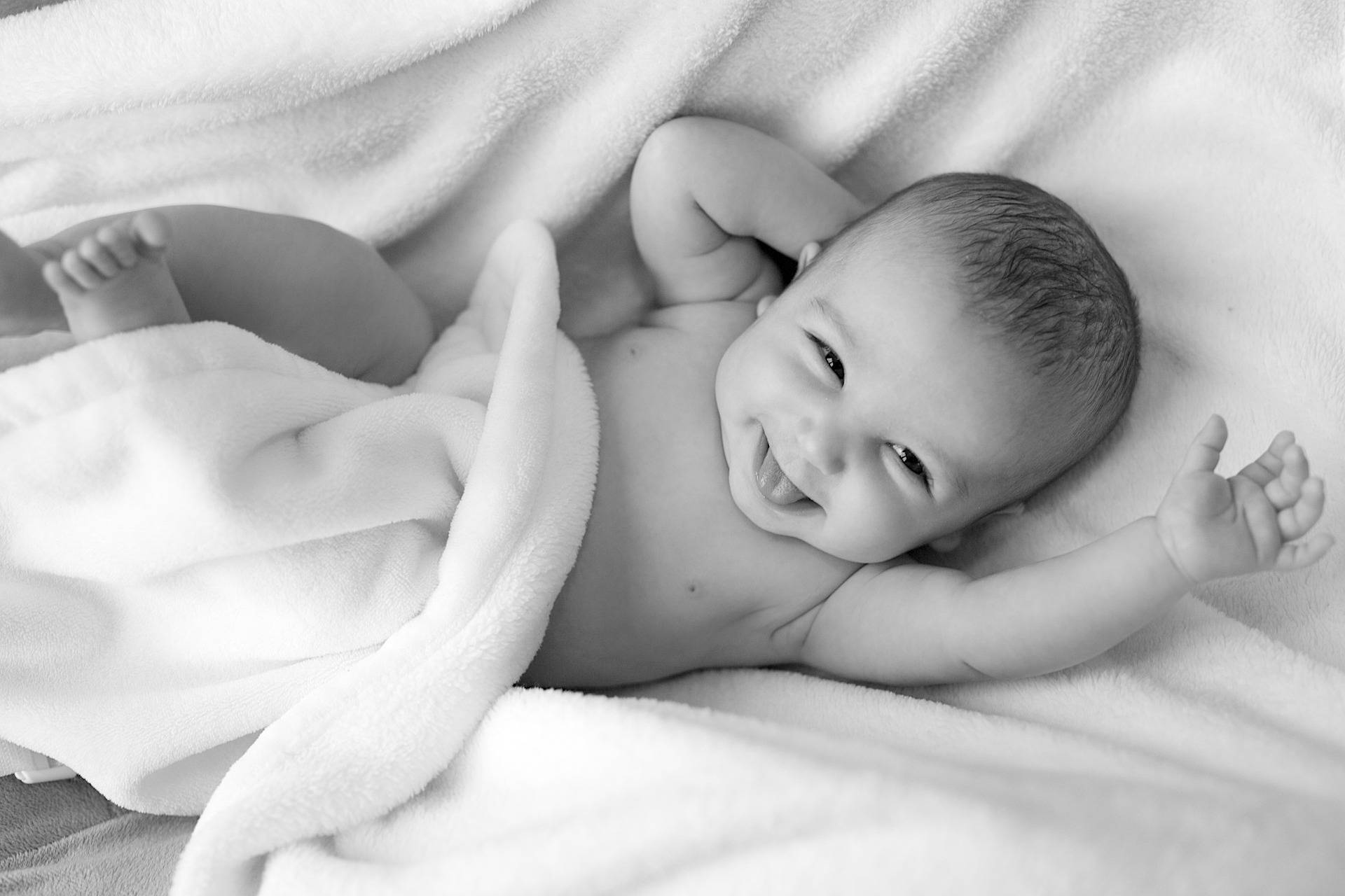 Very Cute Baby 桌布 壁紙 — 一個快樂的嬰兒躺在柔軟的白色毯子上，在溫柔的黑白肖像中明亮地笑著。