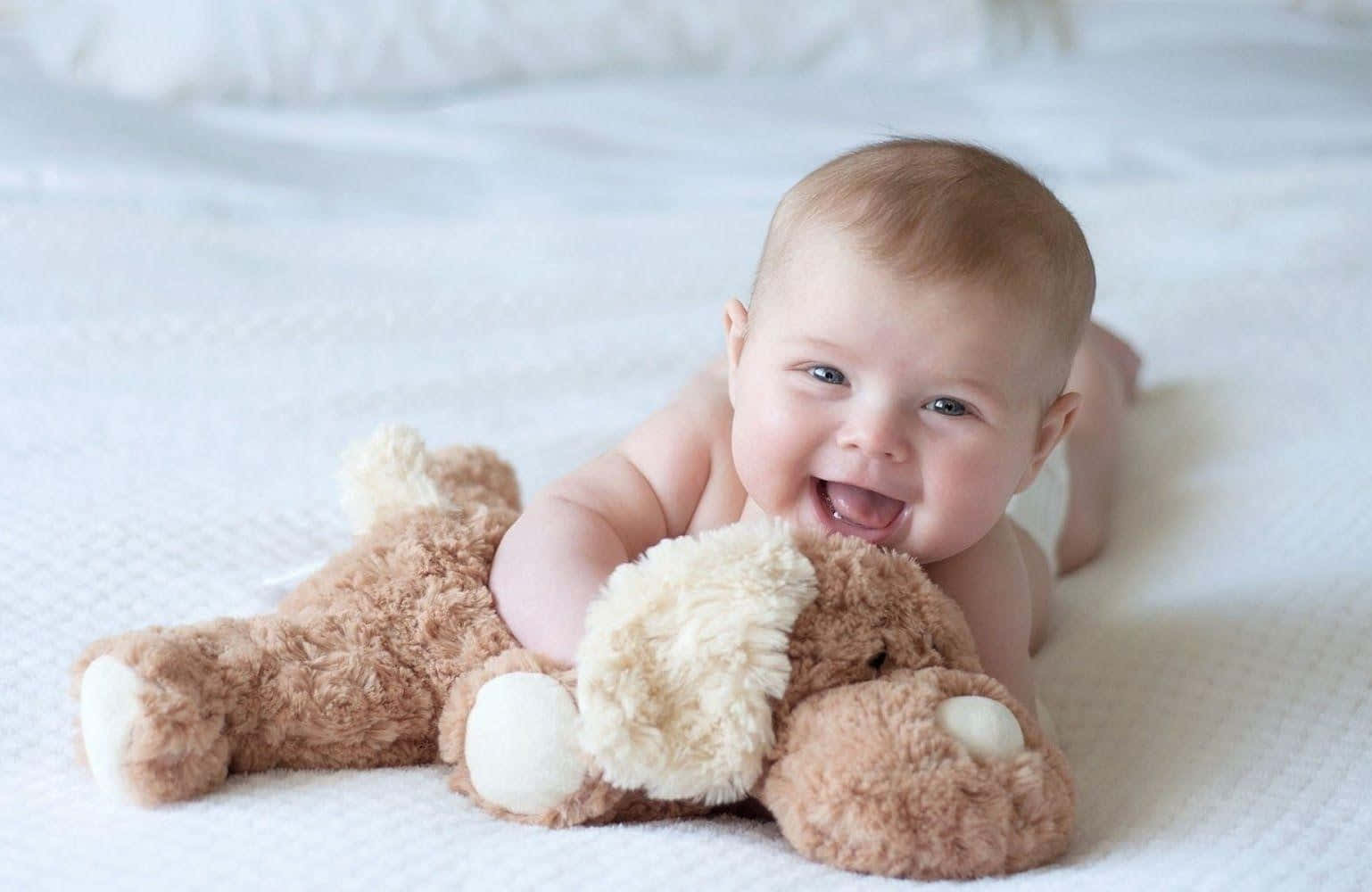 cute baby 桌布 壁紙 — 一個快樂的嬰兒躺在白色床上，抱著一隻有奶油色爪子的棕色泰迪熊，開心地笑著。