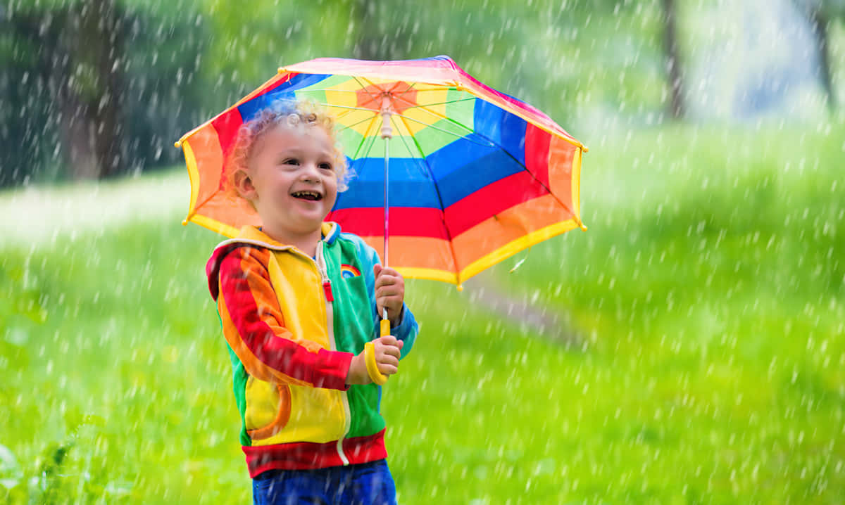 下雨 桌布 壁紙 — 一個快樂的孩子在彩虹傘下大笑，雨水落在翠綠的田野上。