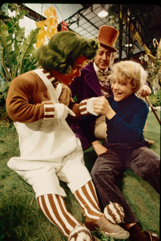 Willy Wonka 桌布 壁紙 — 一個快樂的孩子在玻璃屋頂下的人造草地上與一個綠色頭髮、穿著條紋服裝的角色碰拳。