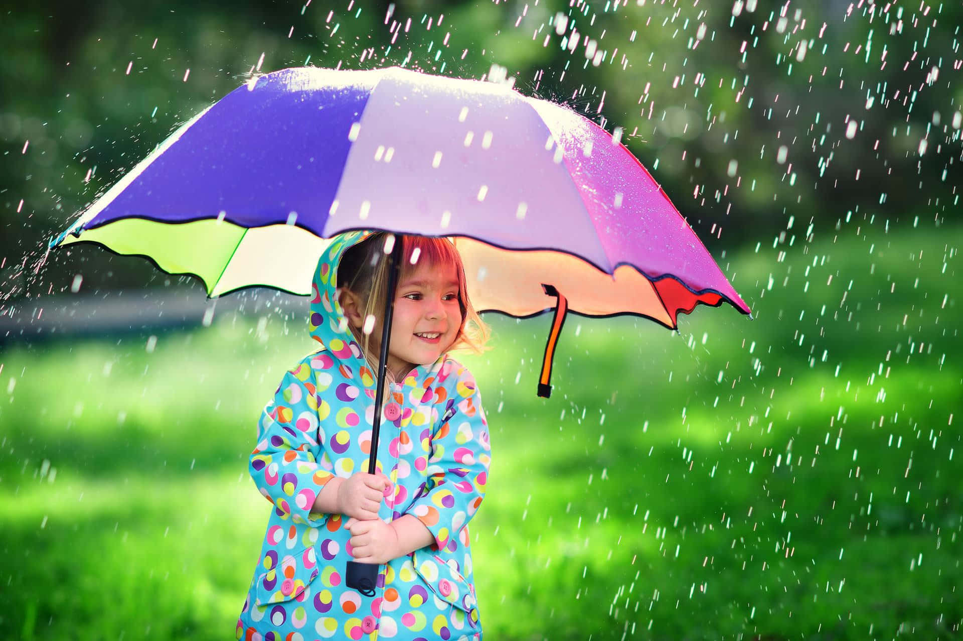 Umbrella 桌布 壁紙 — 一個快樂的孩子穿著波點雨衣，在綠色公園中下雨時微笑著撐著彩虹傘。