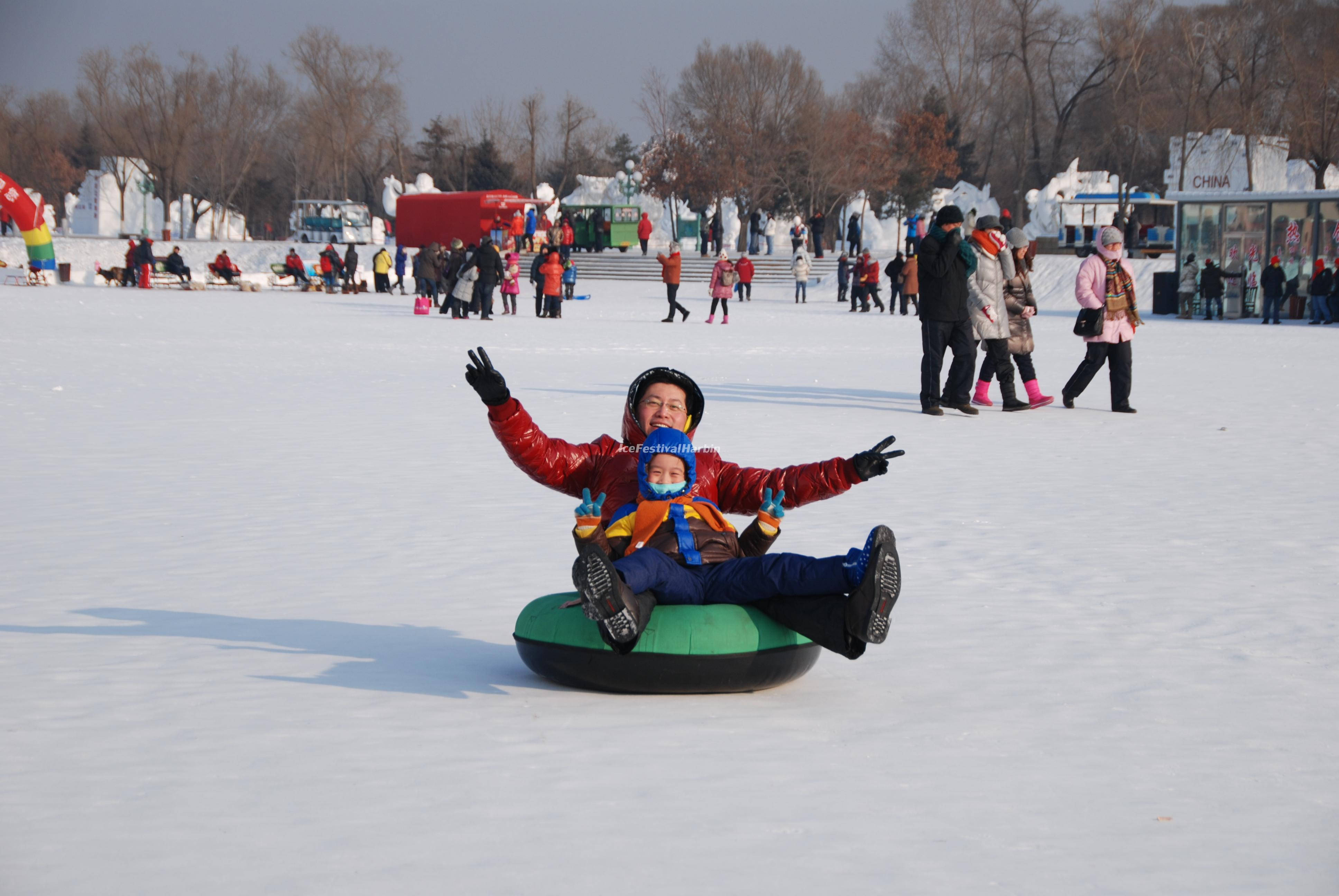 Harbin 桌布 壁紙 — 一個快樂的孩子穿著紅色外套和頭盔，在廣闊的雪地上騎著綠色雪管，背景有光禿禿的樹木和其他人。