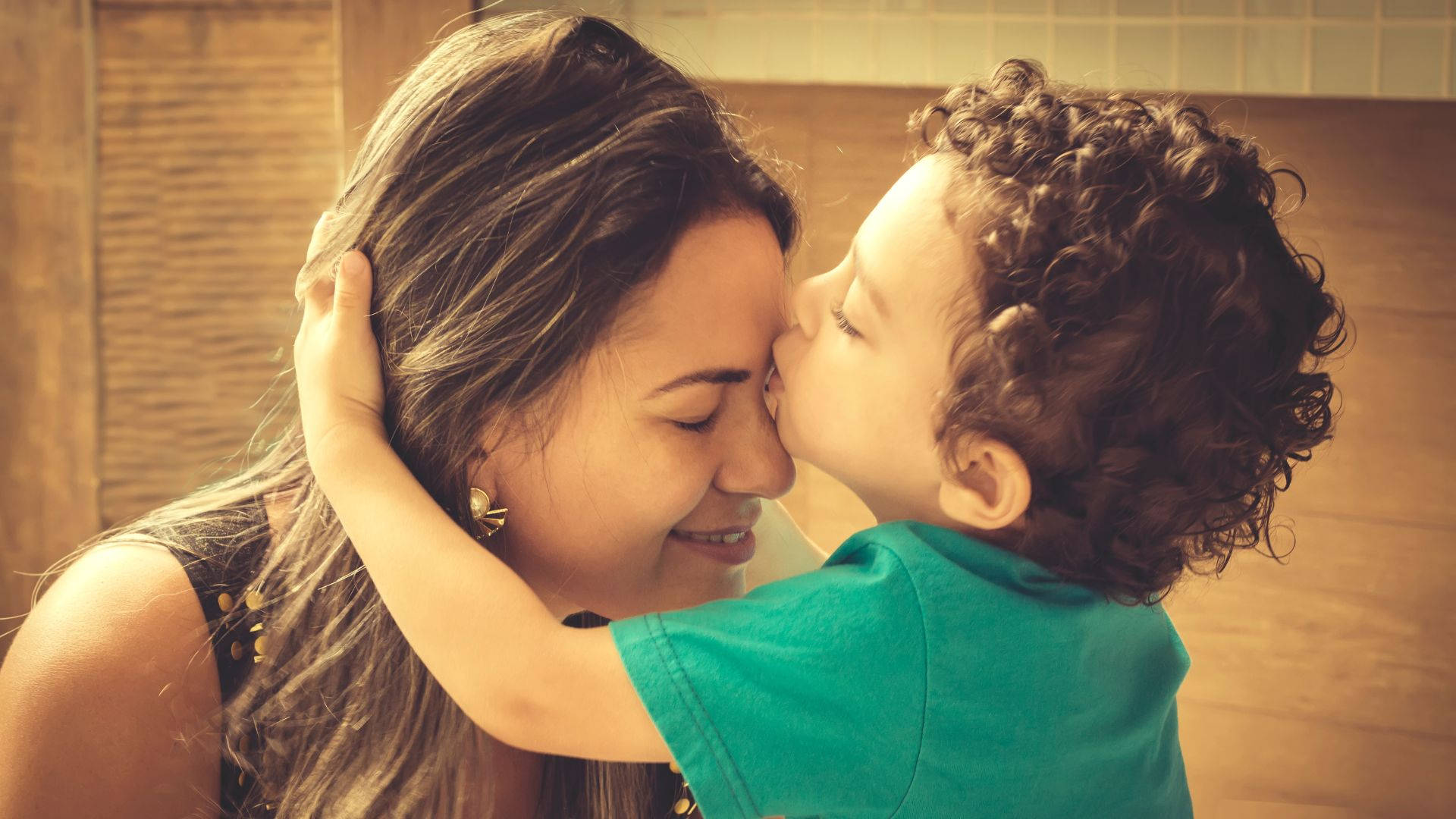 Mom 桌布 壁紙 — 一個快樂的孩子親吻母親的臉頰，母親在陽光灑落的木屋中溫暖地微笑。