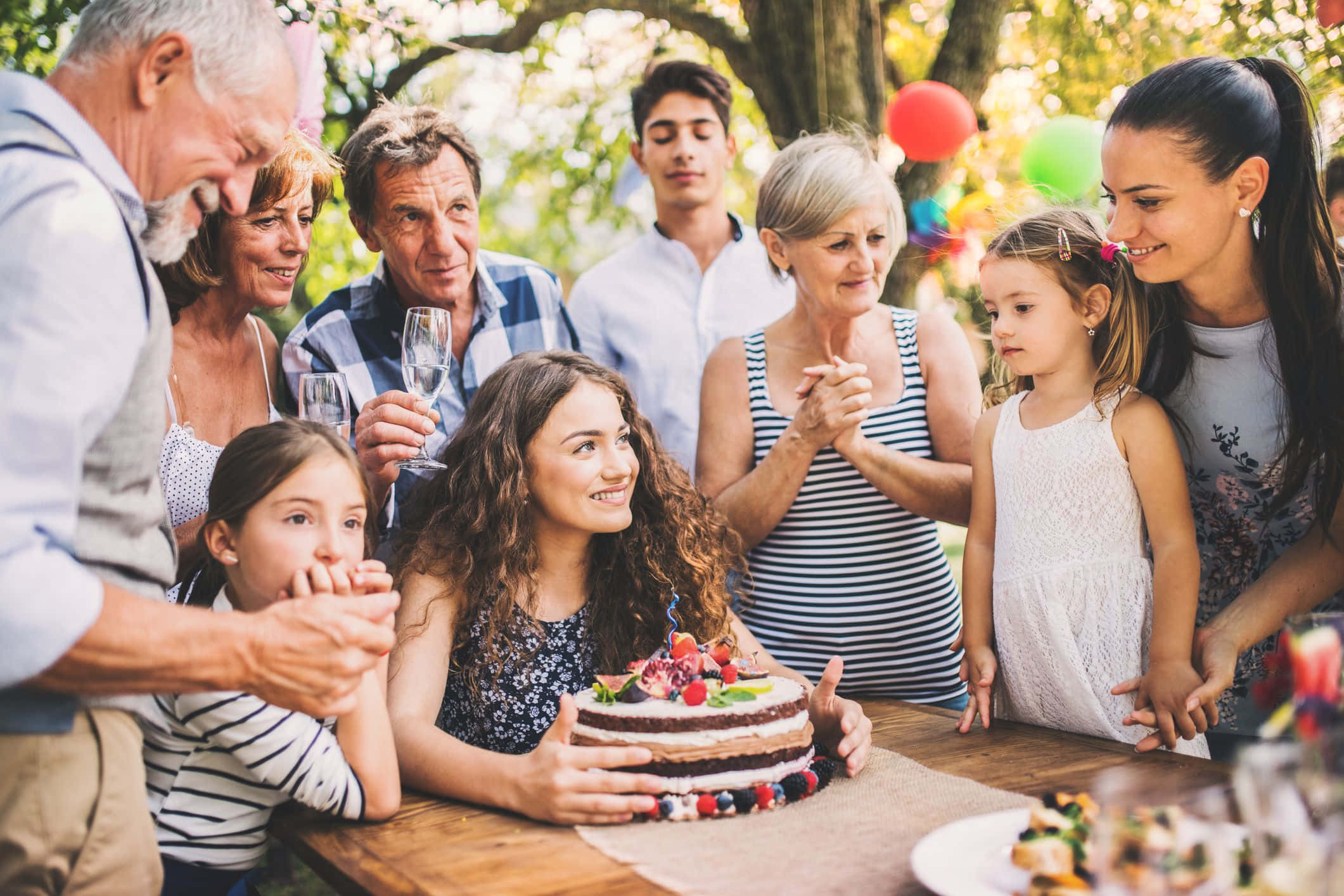 Birthday Celebration 桌布 壁紙 — 一個快樂的家庭在戶外樹下圍繞著生日蛋糕，周圍有色彩繽紛的氣球。