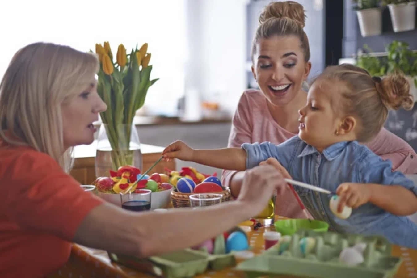 Family 桌布 壁紙 — 一個快樂的家庭在陽光普照的廚房桌上一起繪畫復活蛋，旁邊有鬱金香和色彩繽紛的用品。