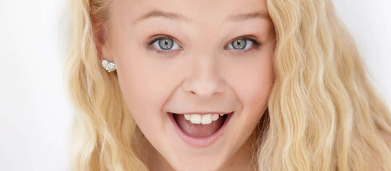 JoJo Siwa 桌布 壁紙 — 一個快樂的小女孩，擁有明亮的藍眼睛和金色捲髮，在純白背景上露出燦爛的笑容。