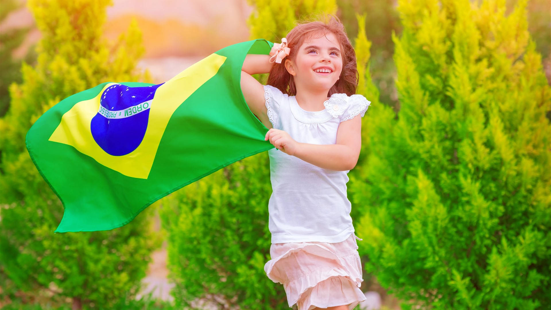 Brazil Flag 桌布 壁紙 — 一個快樂的小女孩在溫暖的陽光下，在充滿活力的綠色灌木叢中自豪地揮舞著巴西國旗。