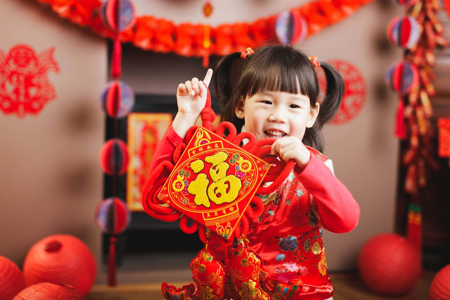 新年 桌布 壁紙 — 一個快樂的小女孩穿著紅色節日服裝，手持一個帶笑臉的裝飾性紅色和金色農曆新年飾物。