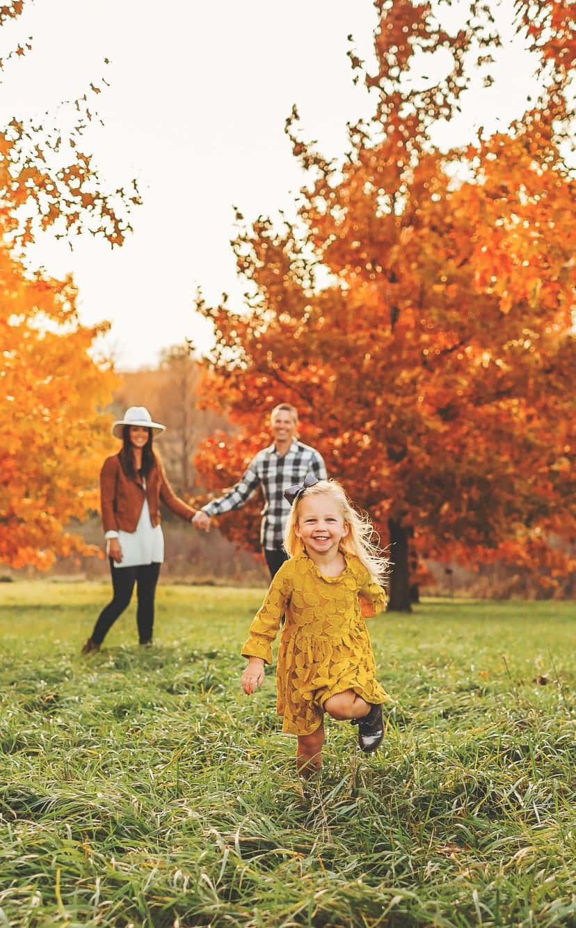Family 桌布 壁紙 — 一個快樂的小女孩穿著黃色連衣裙在草地上奔跑，她的父母在身後手牽手，背景是色彩繽紛的秋樹。