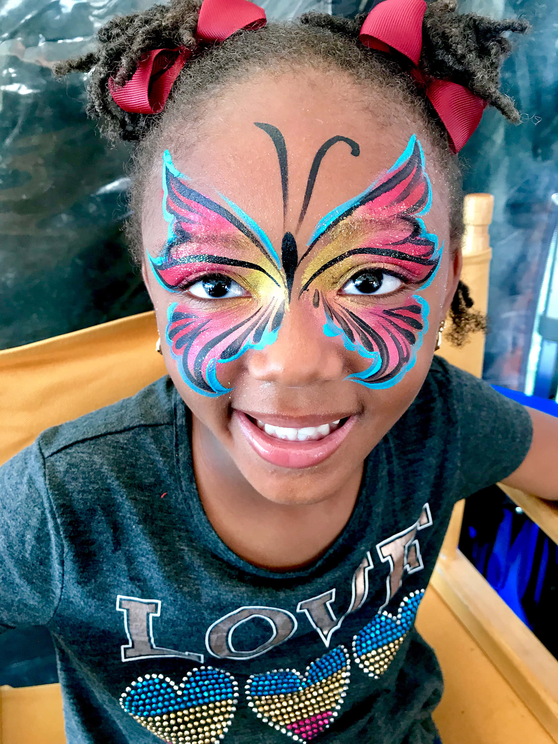 Butterfly Face Painting 桌布 壁紙 — 一個快樂的小女孩臉上畫著鮮豔的蝴蝶圖案，頭髮上繫著紅色蝴蝶結，笑容燦爛。