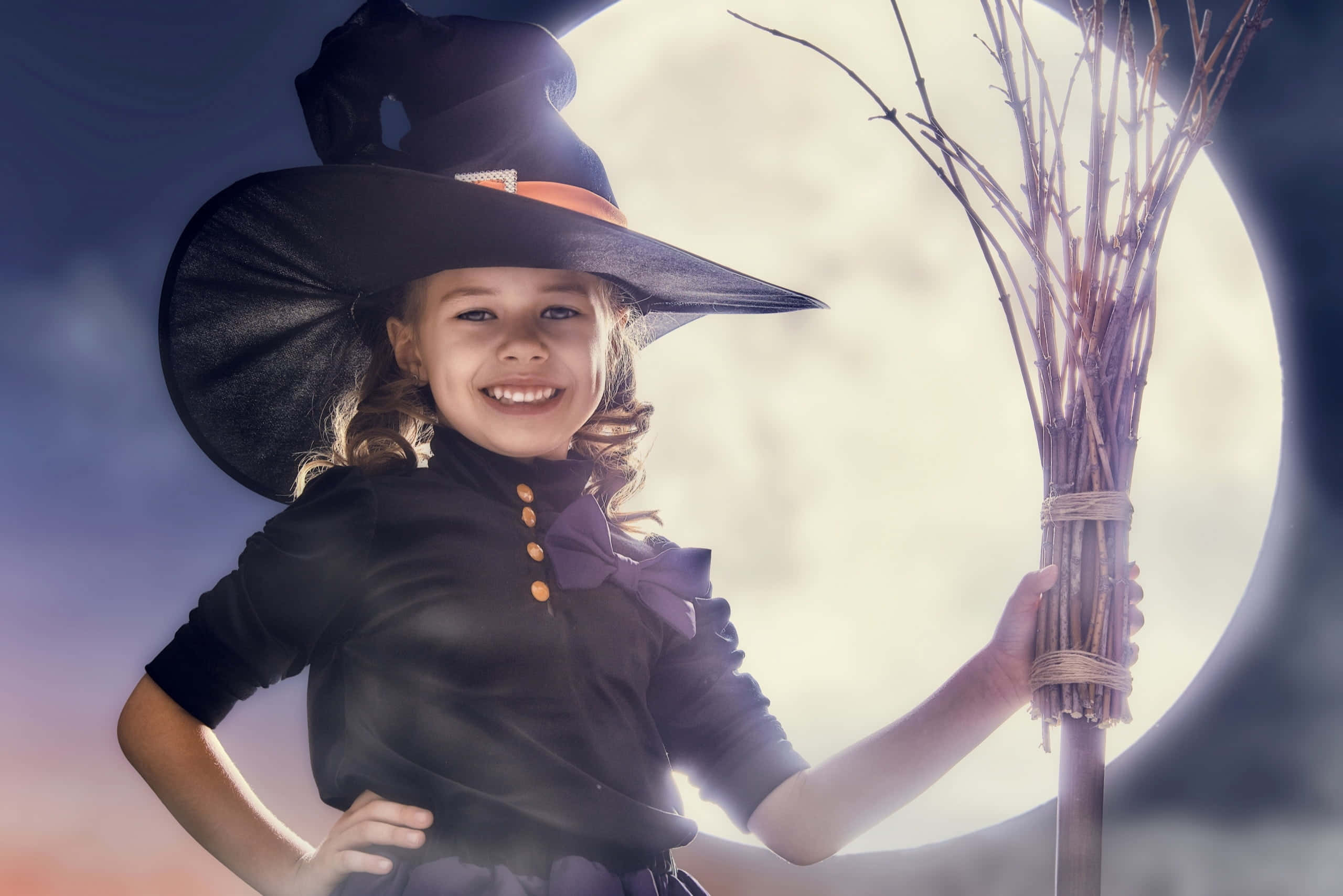 Halloween Costume 桌布 壁紙 — 一個快樂的小女巫，戴著黑色帽子和穿著黑色裙子，在發光的滿月背景下微笑著拿著掃帚。