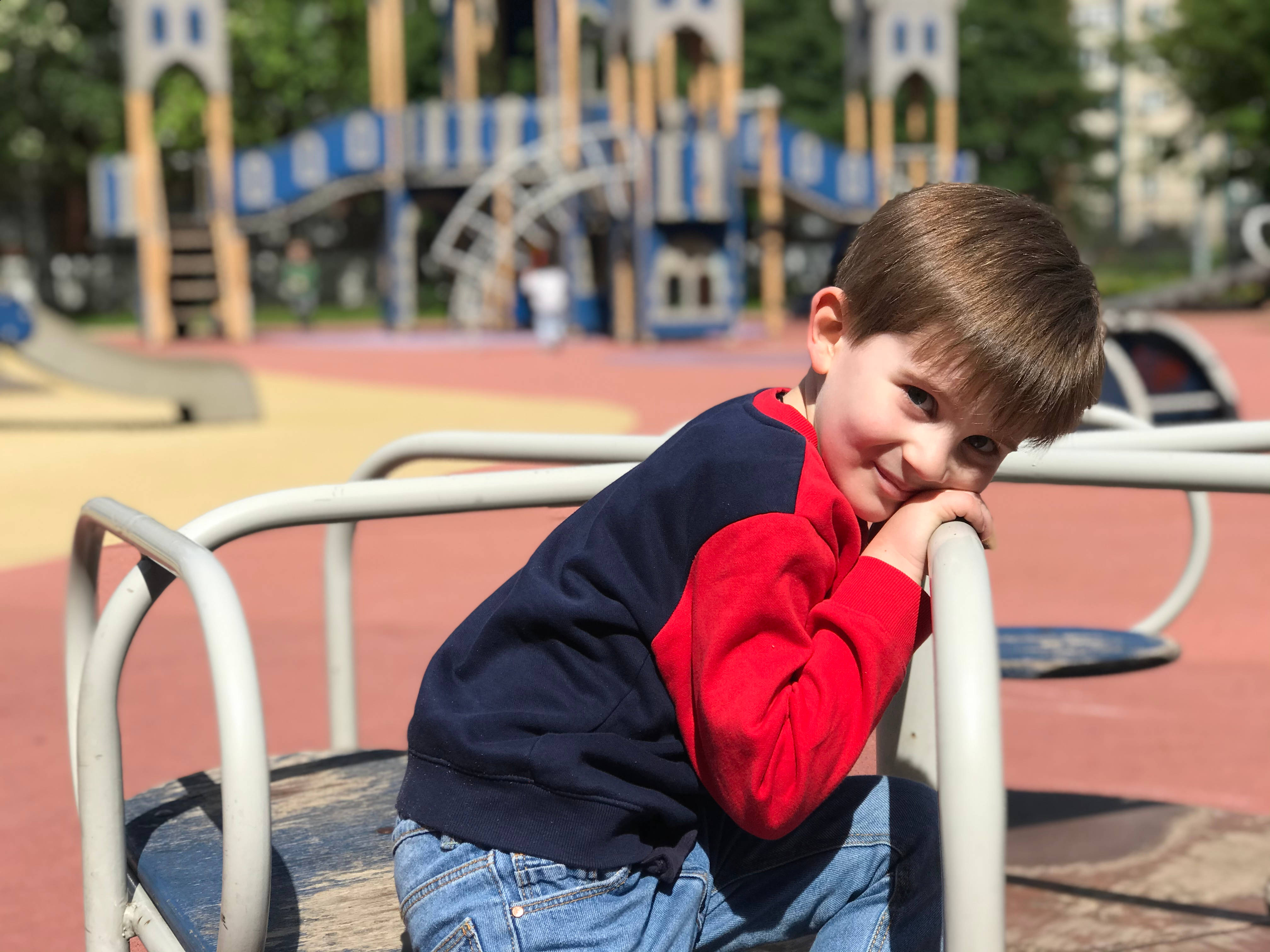 Playground 桌布 壁紙 — 一個快樂的小男孩穿著紅色和深藍色運動衫，在明亮的陽光下靠在遊樂場的旋轉木馬上微笑。
