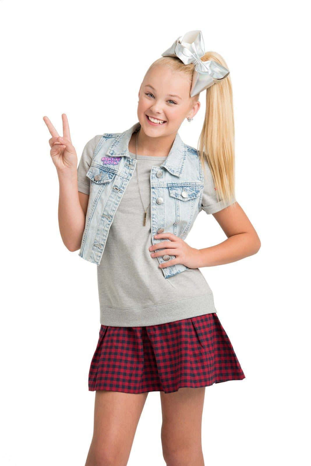 JoJo Siwa 桌布 壁紙 — 一個快樂的年輕女孩，金髮紮成馬尾辮，穿著牛仔背心和格子裙，微笑著比出和平手勢。