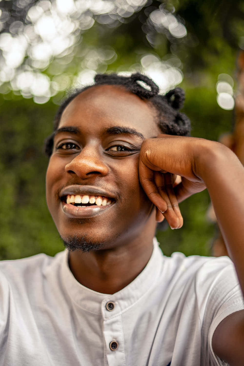 Smile 桌布 壁紙 — 一個快樂的年輕男子，編著辮子，手托下巴，在茂密的綠色散景背景下燦爛地微笑。