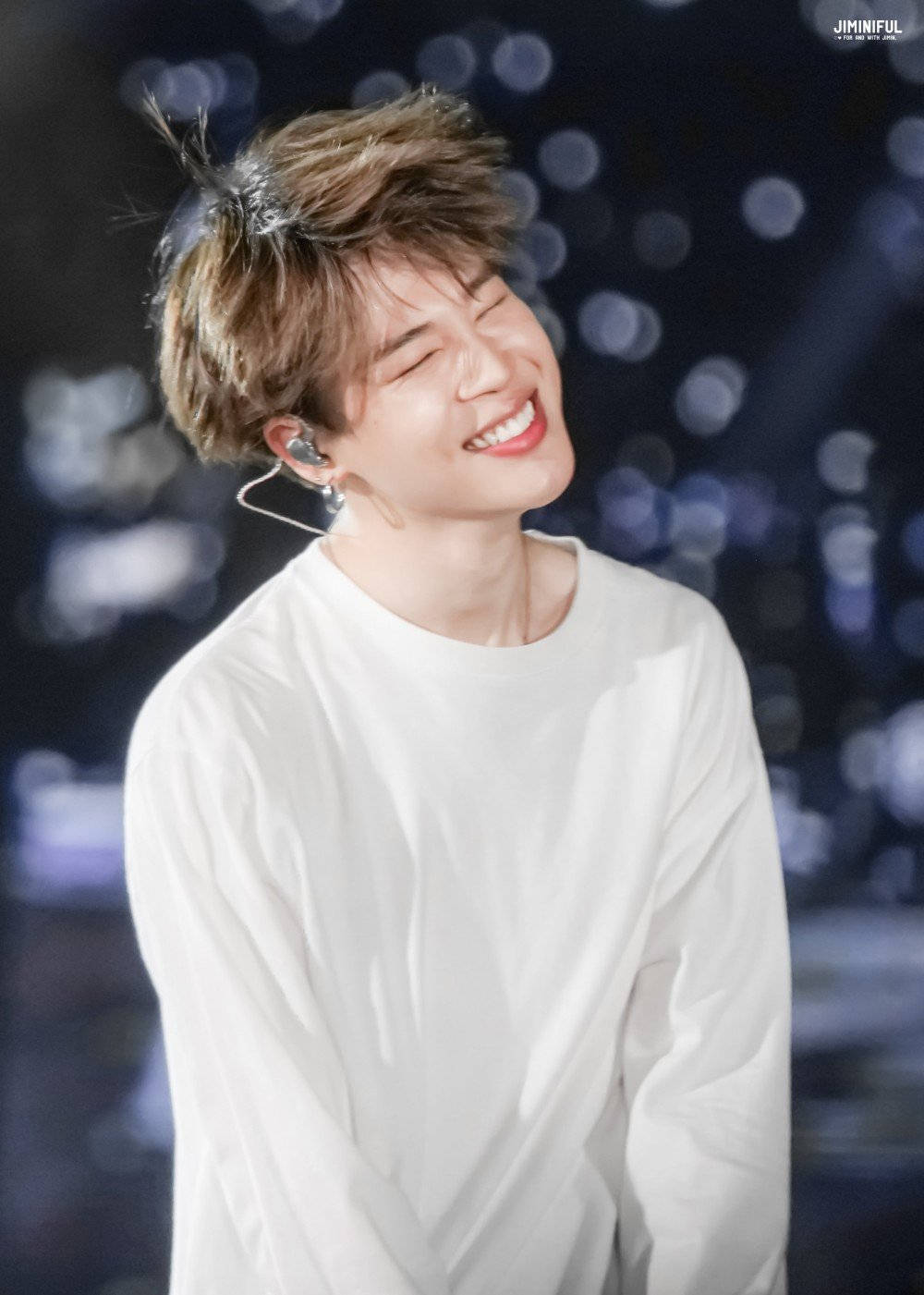BTS 智旻 桌布 壁紙 — 一個快樂的年輕男子，頭髮蓬鬆，戴著耳機，在模糊的舞台背景下閃耀著光芒。