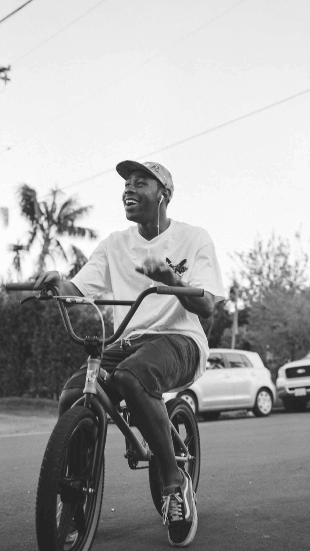造物主泰勒 桌布 壁紙 — 一個快樂的年輕男子騎著BMX自行車沿著一條有棕櫚樹和汽車背景的街道行駛，黑白照片。