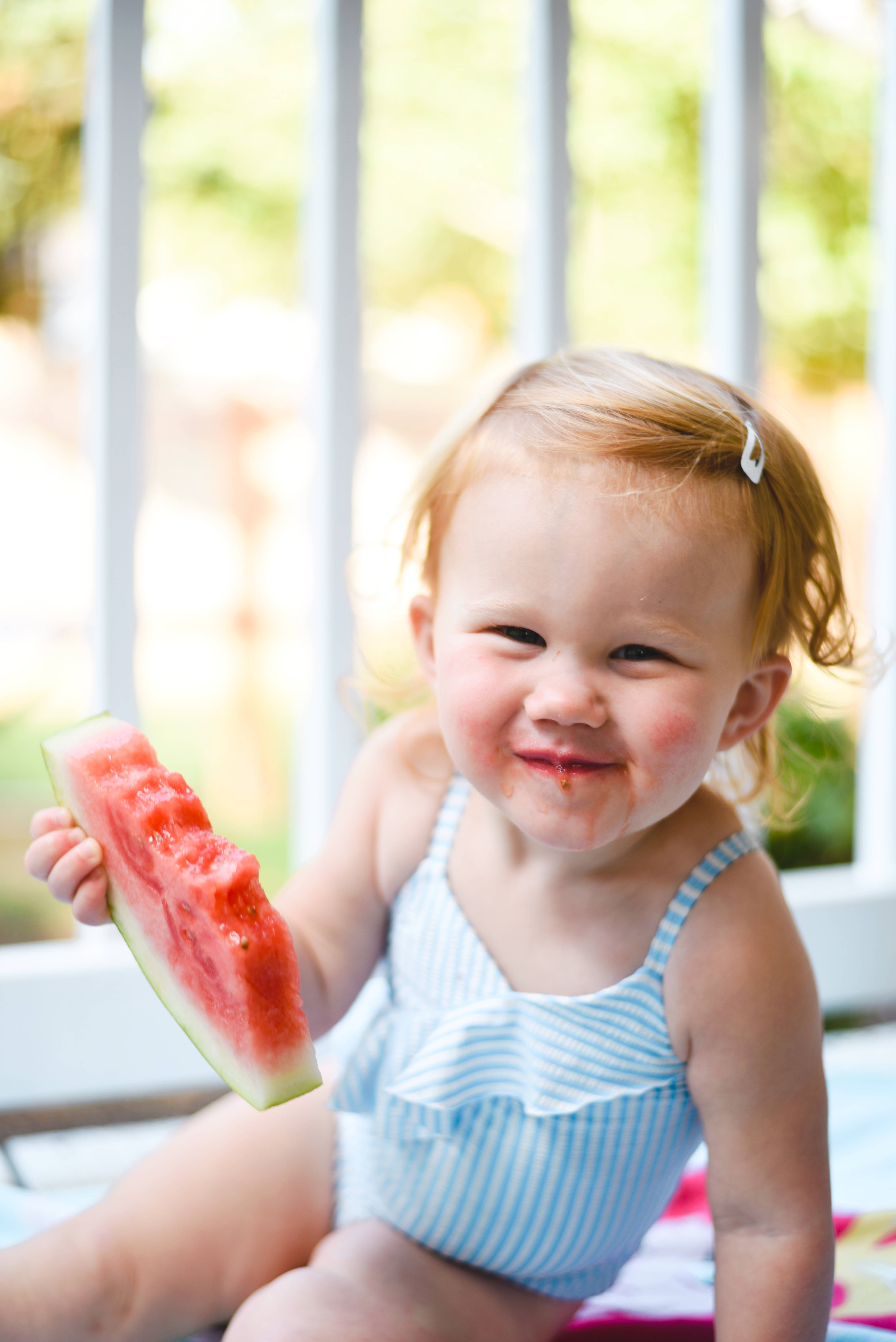 Watermelon 桌布 壁紙 — 一個快樂的幼兒，臉頰髒兮兮，在陽光明媚的門廊上開心地吃著多汁的西瓜片。