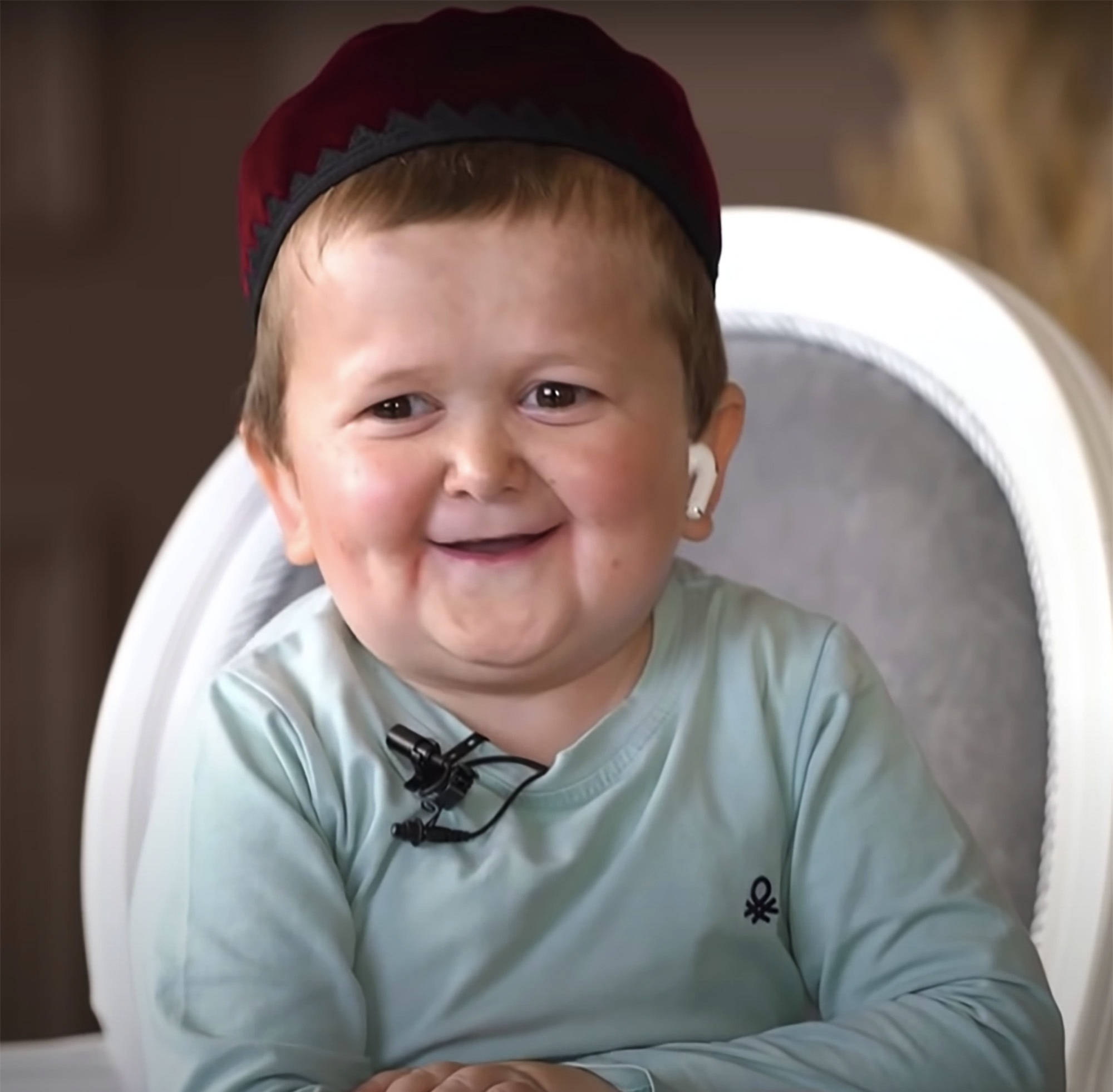 Smile 桌布 壁紙 — 一個快樂的幼兒戴著栗色帽子和淺藍色襯衫，坐在白色椅子上燦爛地微笑。