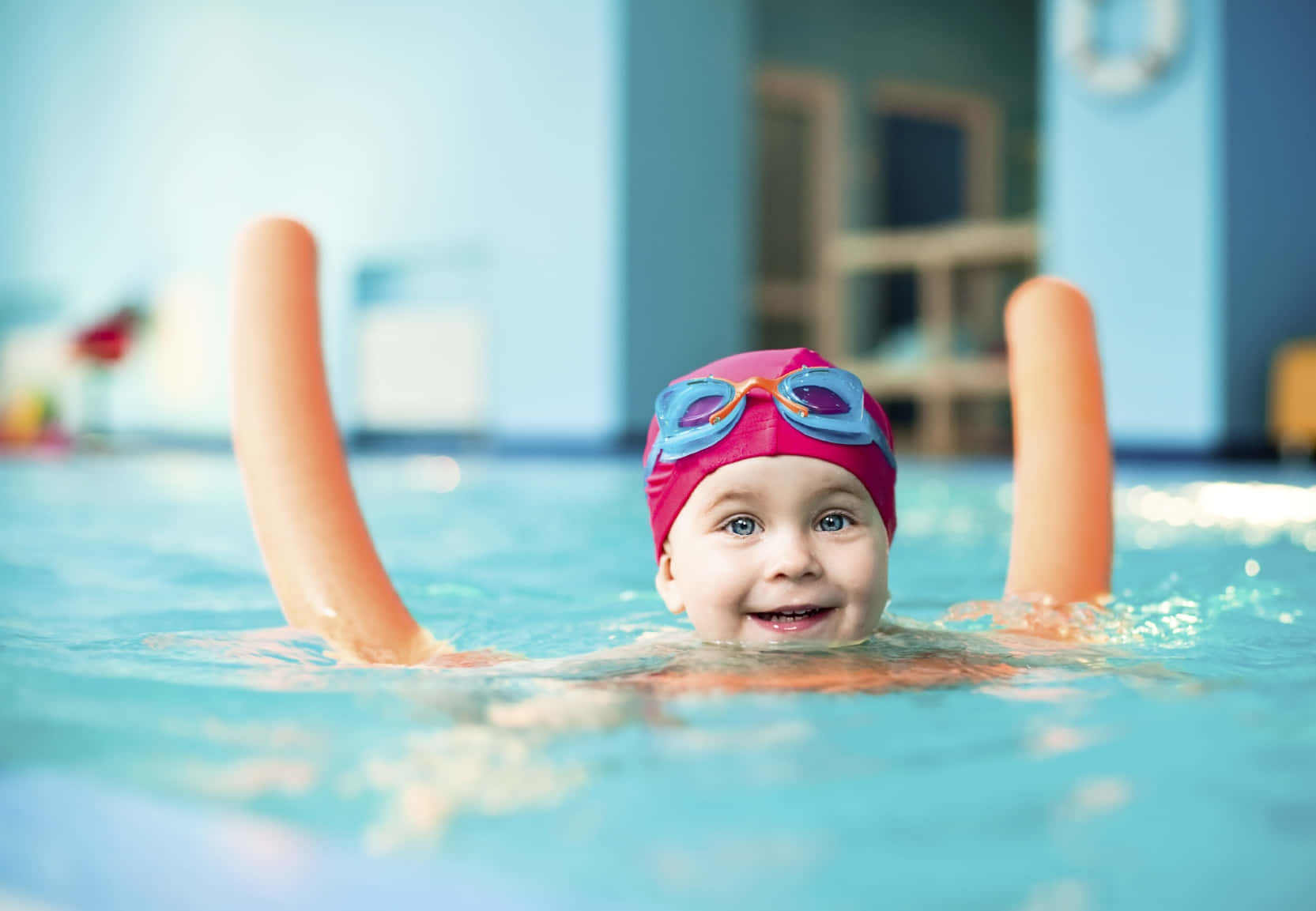 Swimming 桌布 壁紙 — 一個快樂的幼兒戴著粉紅色泳帽和泳鏡，在明亮的室內游泳池中帶著橙色浮水袖游泳，面帶微笑。