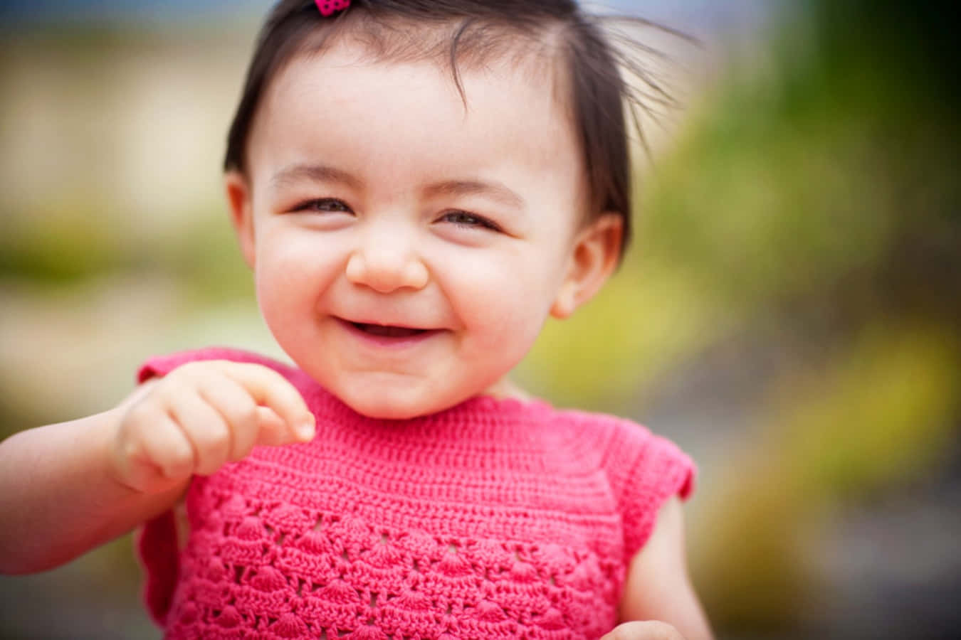 toddler 桌布 壁紙 — 一個快樂的幼兒穿著亮粉紅色鉤針編織上衣，露出燦爛的笑容，頑皮地指向前方。
