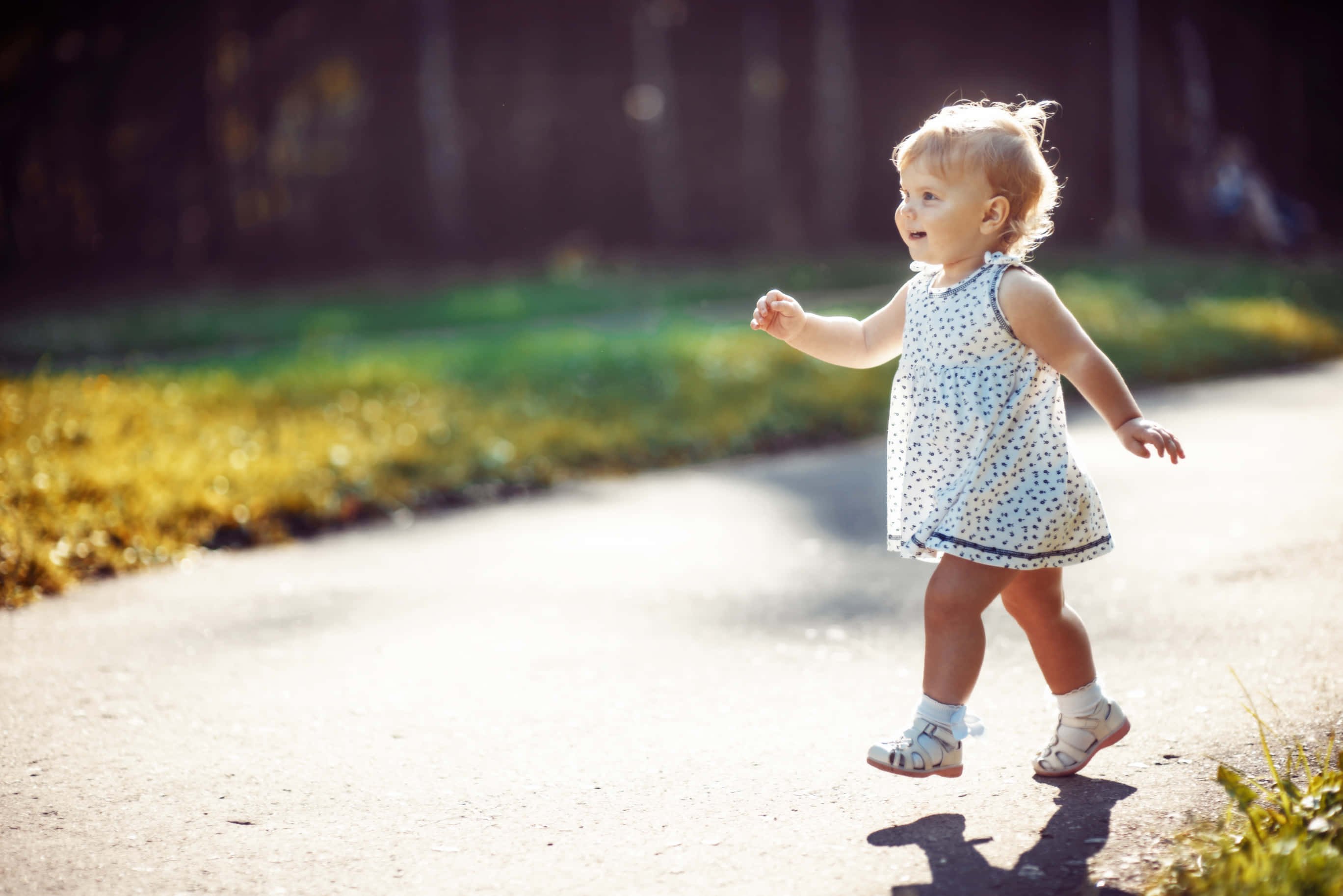 Walking 桌布 壁紙 — 一個快樂的幼兒穿著圓點裙，自信地走在陽光明媚的小路上，背景是柔焦的草地和樹木。