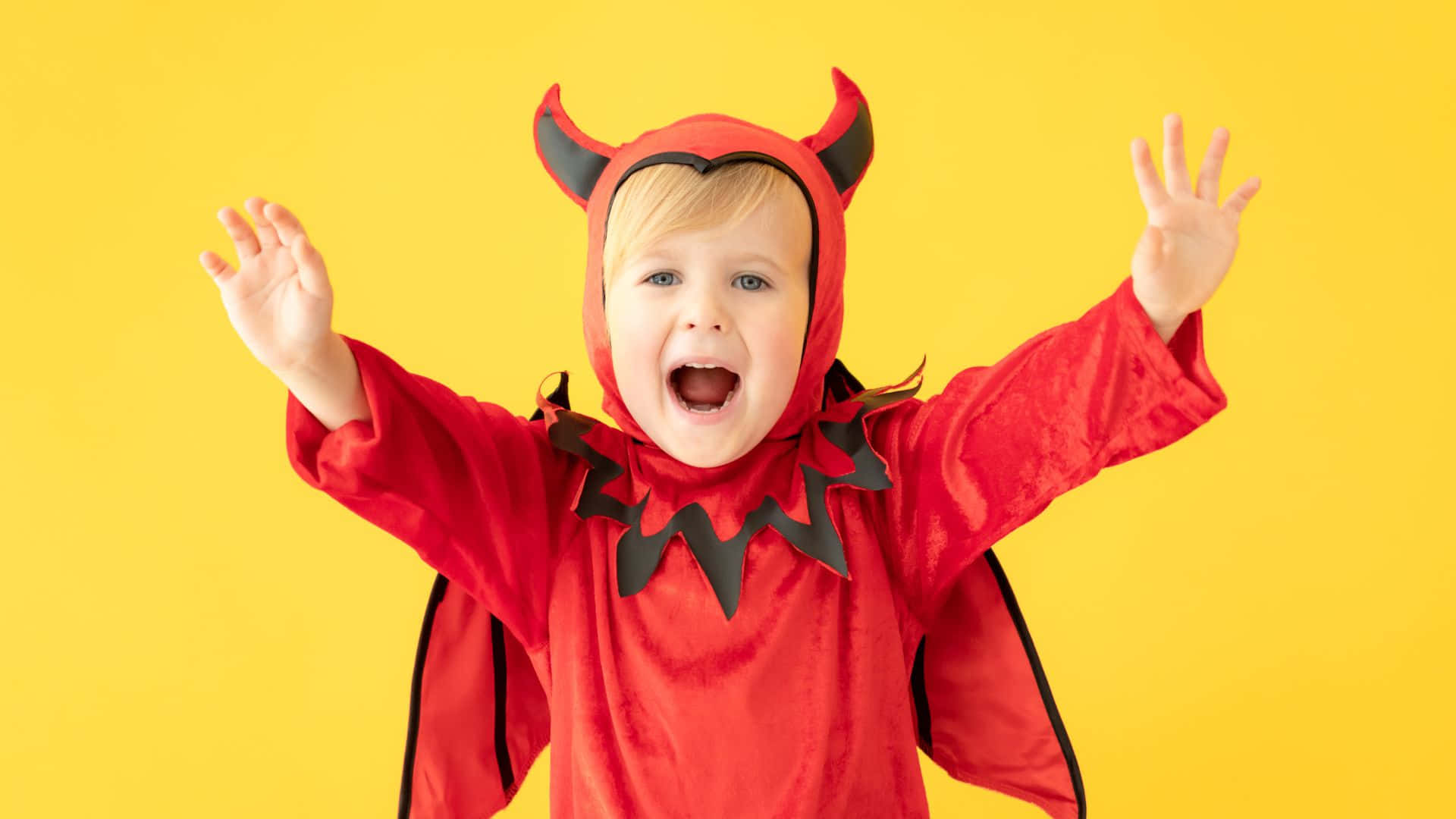 Halloween Costumes 桌布 壁紙 — 一個快樂的幼兒穿著紅色魔鬼服裝，有黑色角和翅膀，在明亮的黃色背景前舉起雙臂，笑容燦爛。