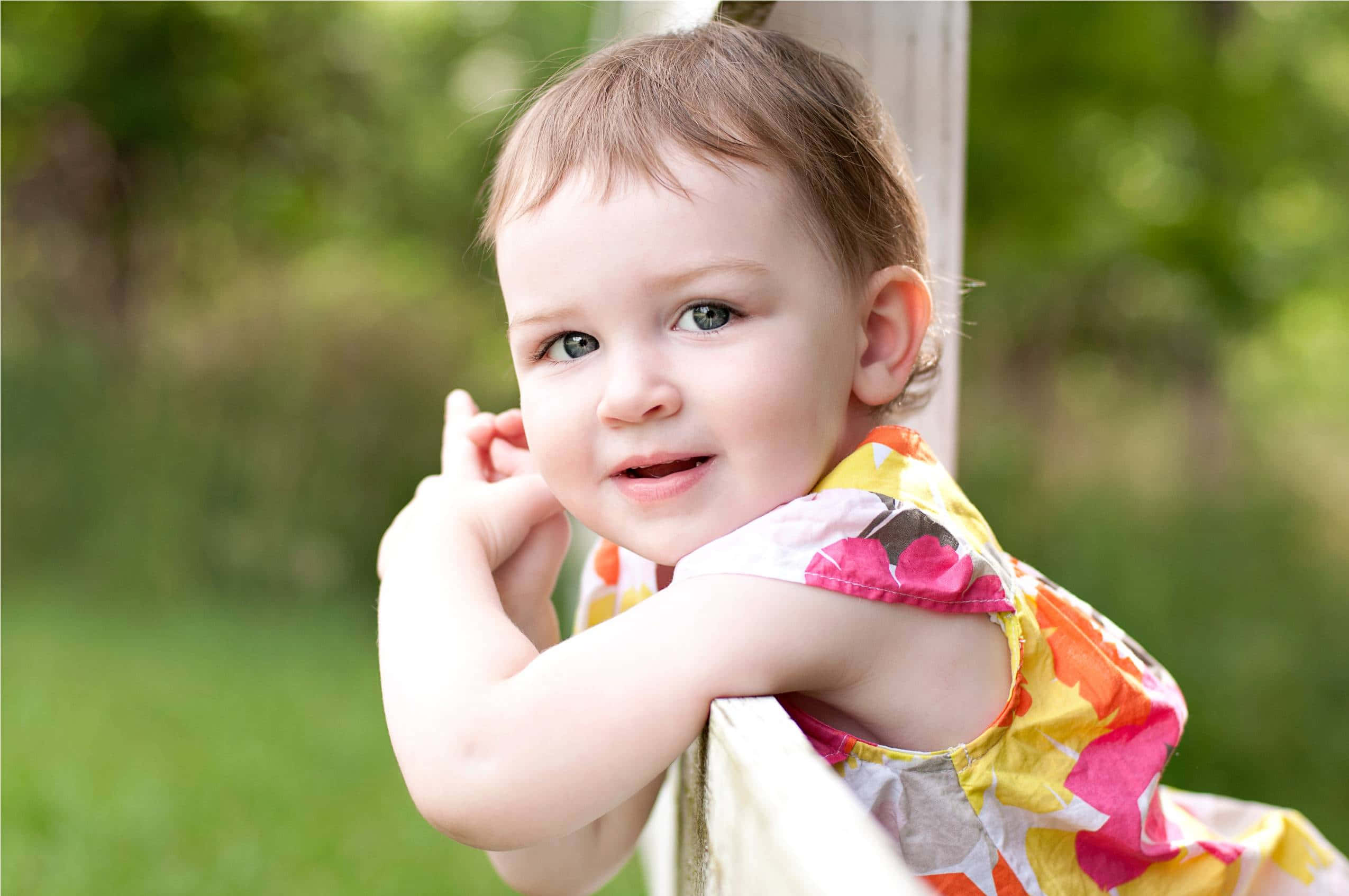 cute baby 桌布 壁紙 — 一個快樂的幼兒穿著色彩繽紛的花裙，靠在木柱上，背景是明亮的綠色模糊花園。