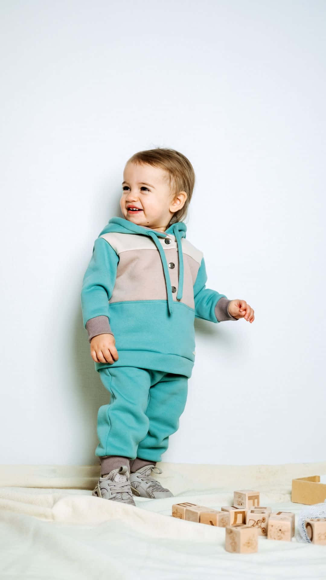 Cute Boy 桌布 壁紙 — 一個快樂的幼兒穿著青綠色和米色連帽衫，在白色牆壁前的柔軟毯子上微笑著站在木積木旁邊。