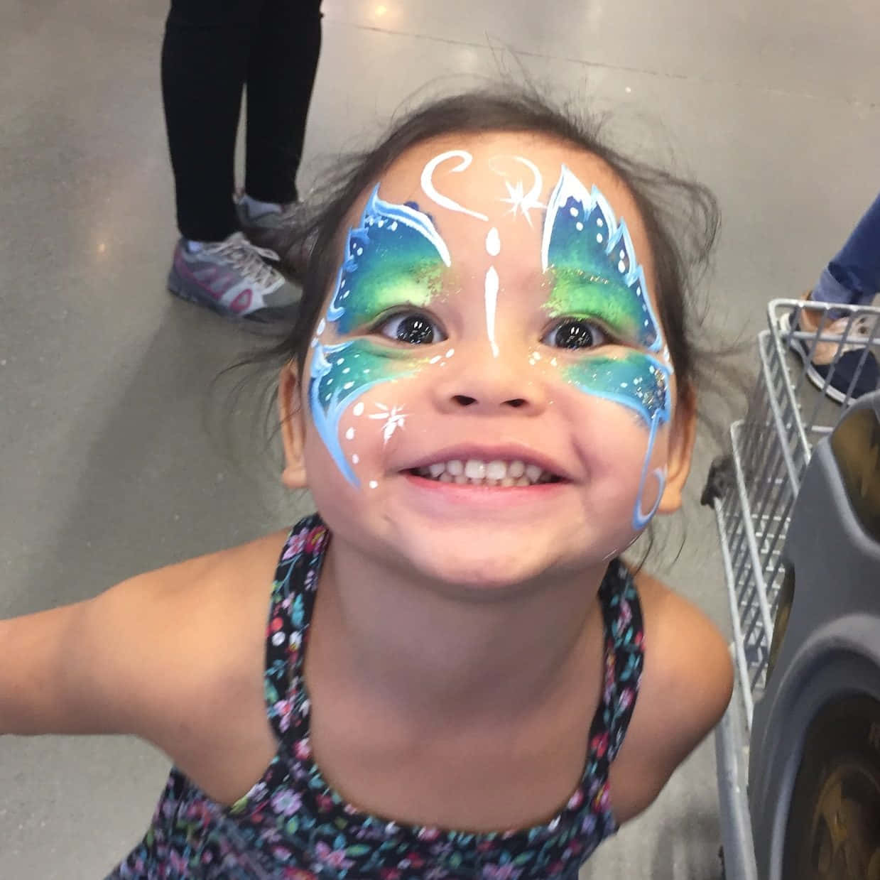 Butterfly Face Painting 桌布 壁紙 — 一個快樂的幼兒臉上畫著鮮豔的藍、綠、白色蝴蝶彩繪，在購物中心對著鏡頭微笑。