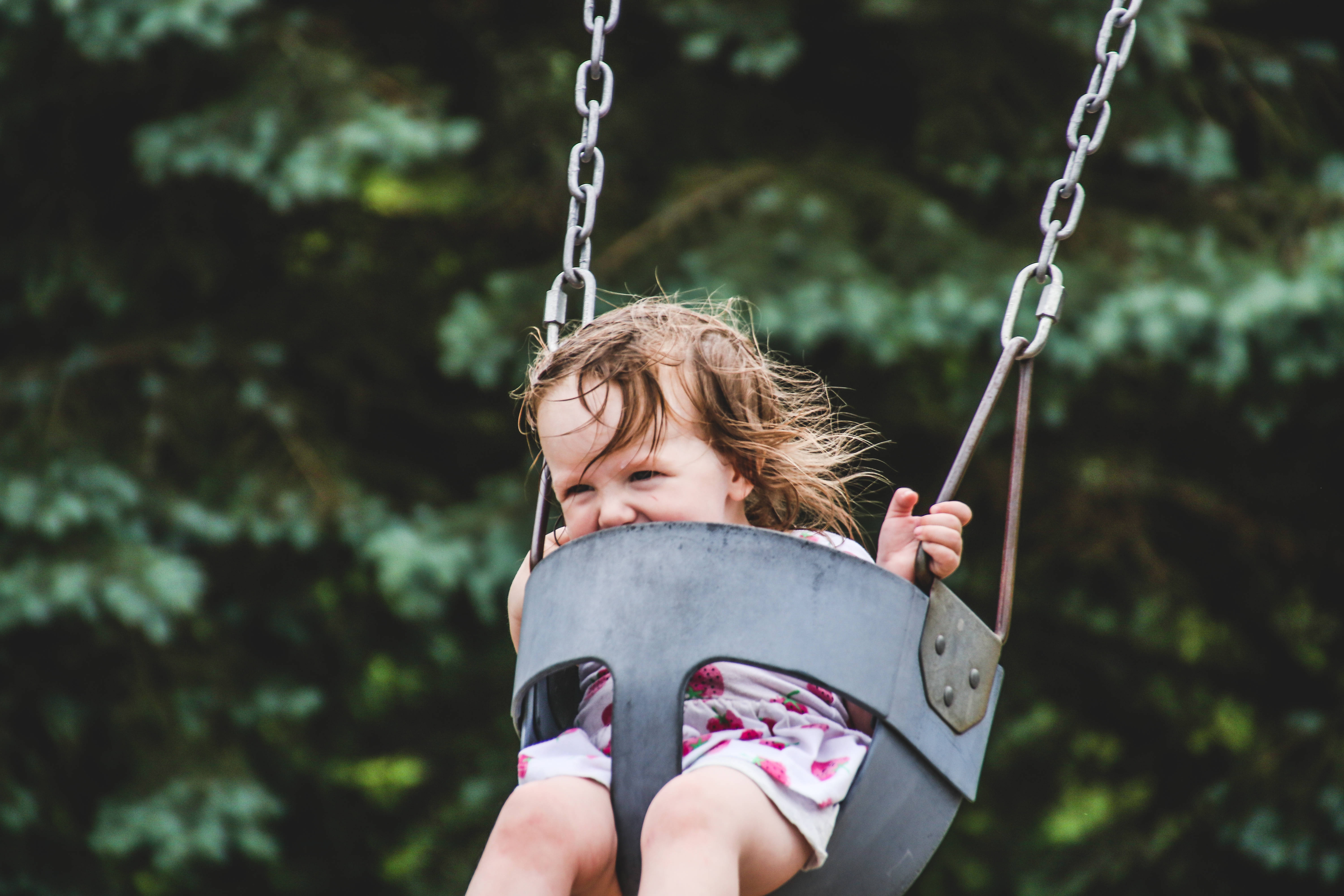 Playground 桌布 壁紙 — 一個快樂的幼兒頭髮蓬亂，在茂密綠樹的背景下盪著金屬鞦韆大笑。