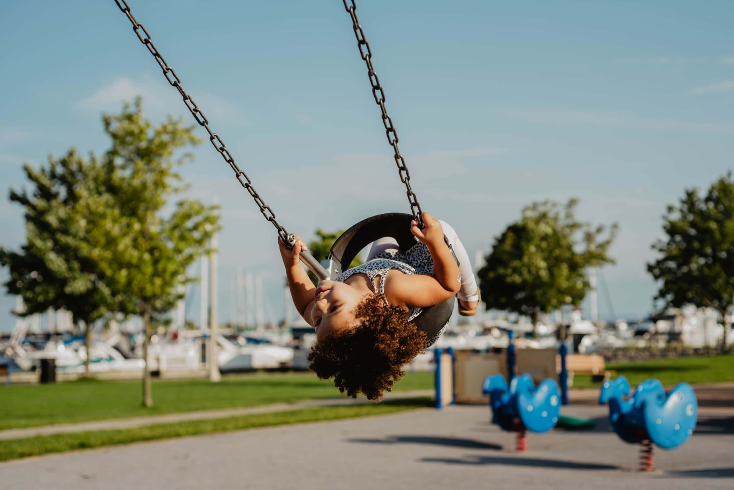 Outdoor Activities 桌布 壁紙 — 一個快樂的捲髮小孩在晴朗藍天下，以綠樹和停泊船隻為背景，高高地盪著鞦韆。
