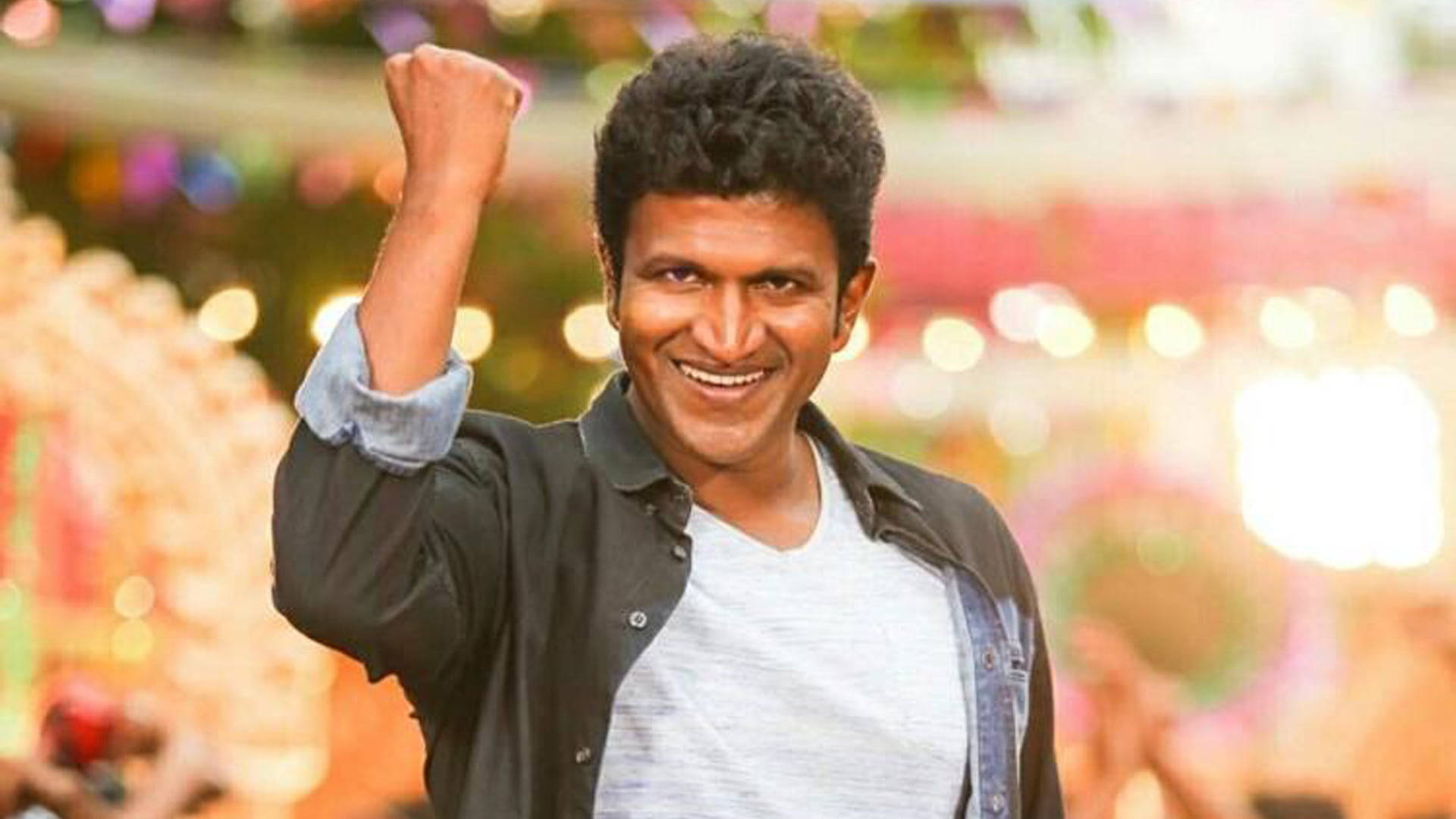 Puneeth Rajkumar 桌布 壁紙 — 一個快樂的捲髮男子在色彩繽紛的散景背景下舉起拳頭慶祝勝利。