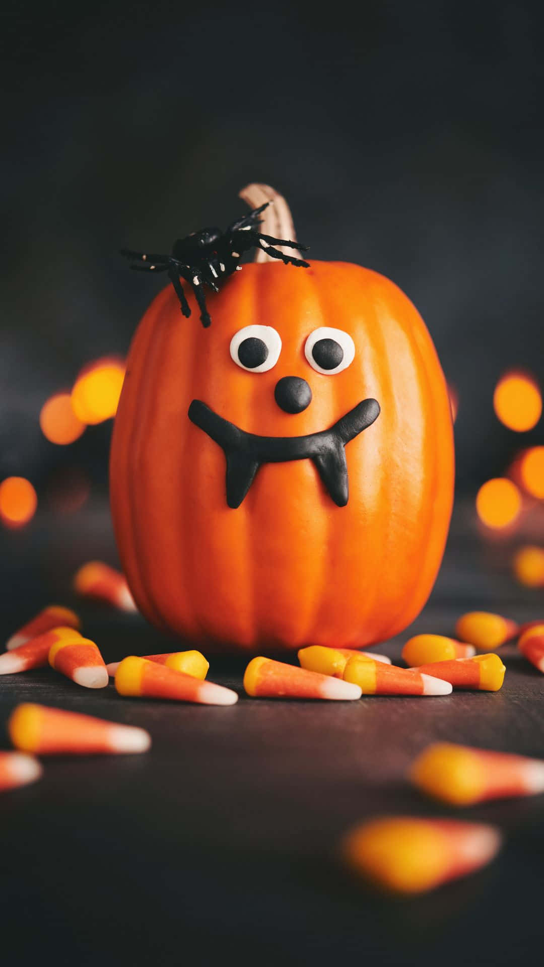 Cute Pumpkin 桌布 壁紙 — 一個快樂的橙色南瓜，帶有咕嚕眼和黑色糖果尖牙，坐在黑暗的表面上，周圍散落著糖果玉米。