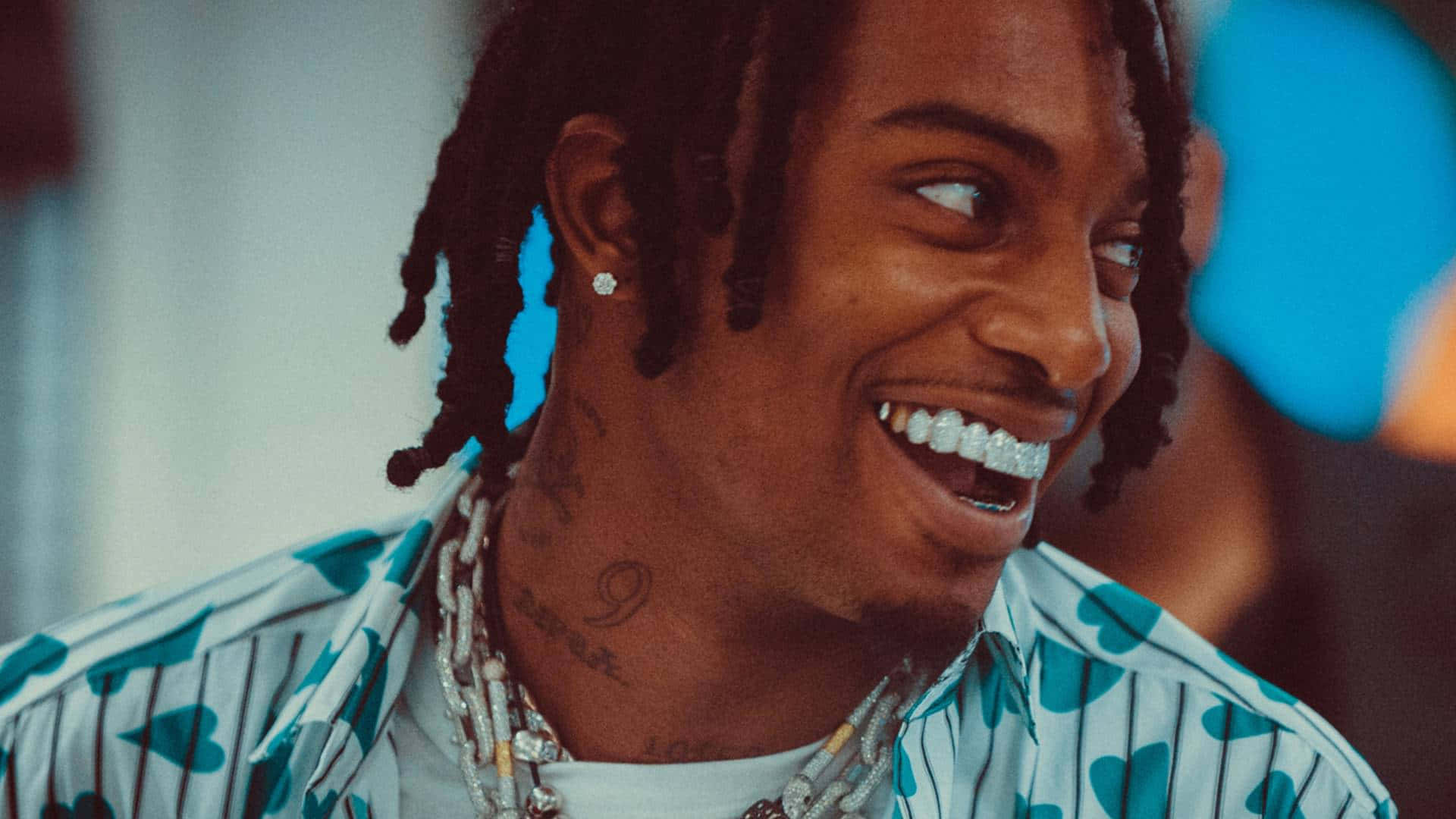 Playboi Carti HD 桌布 壁紙 — 一個快樂的男人，留著辮子頭和鑽石牙套，穿著心形圖案襯衫，開懷大笑。