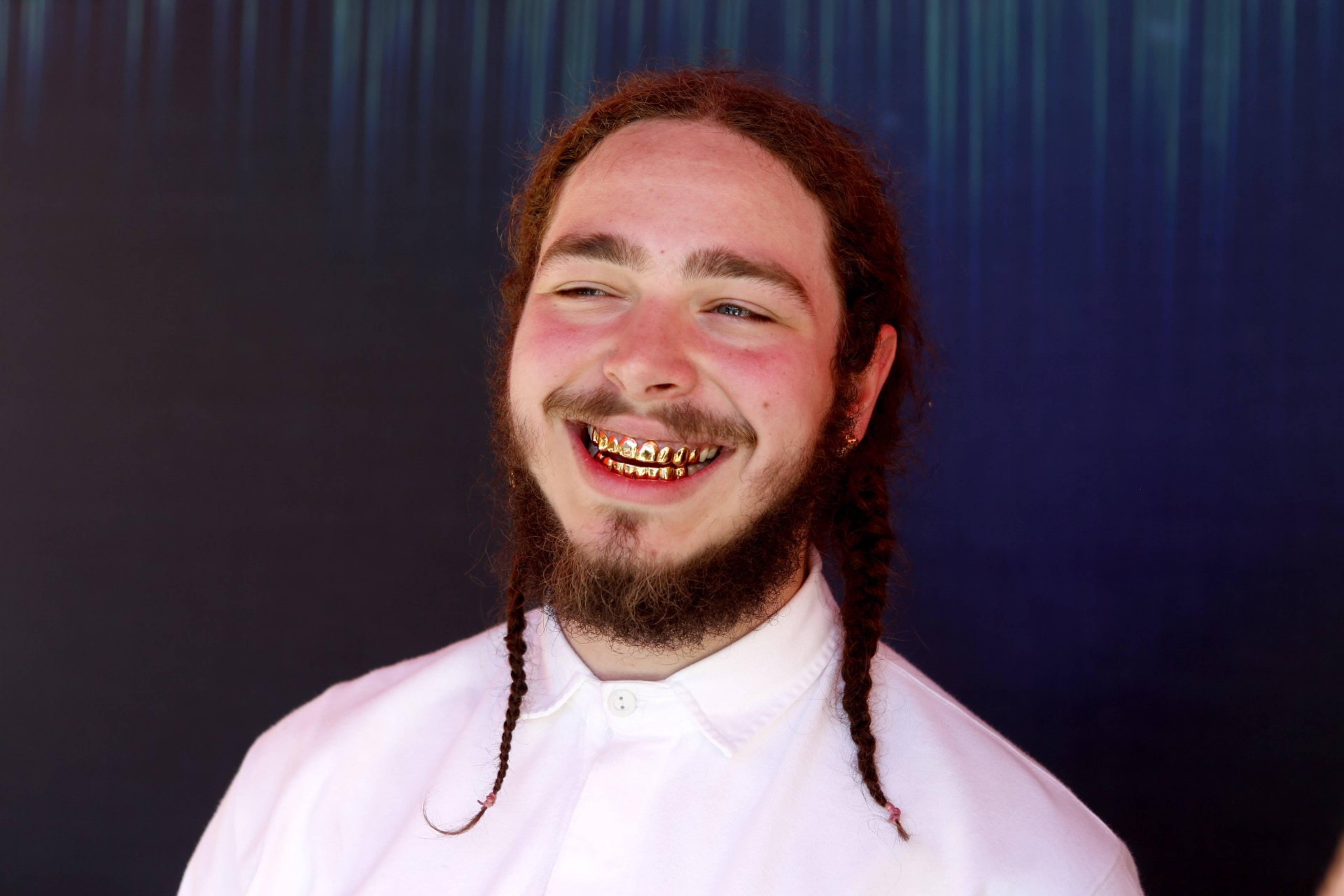 Post Malone 桌布 壁紙 — 一個快樂的男人，辮子頭髮和金色牙套，在深藍色窗簾背景下燦爛地笑。