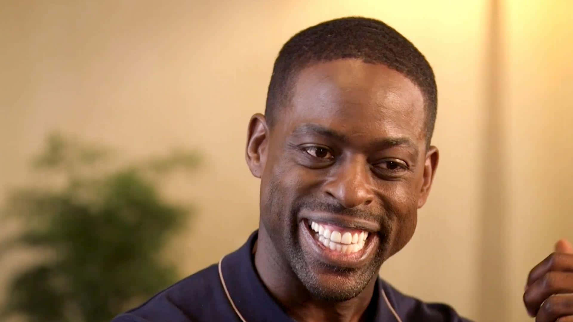 Sterling K Brown 桌布 壁紙 — 一個快樂的男人，露出燦爛的笑容和明亮的眼睛，在溫暖燈光的室內環境中直視鏡頭。