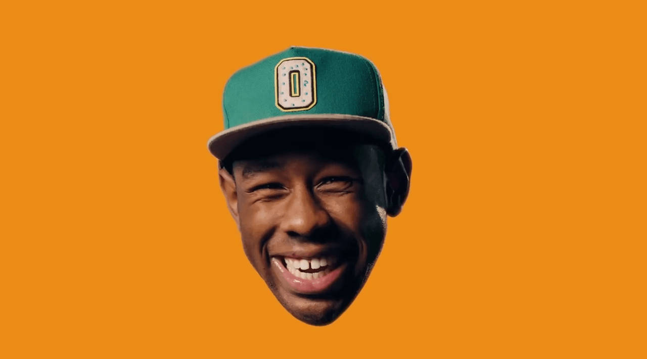 tyler 桌布 壁紙 — 一個快樂的男人戴著帶有'O'標誌的綠色帽子，在純橙色背景上燦爛地微笑。