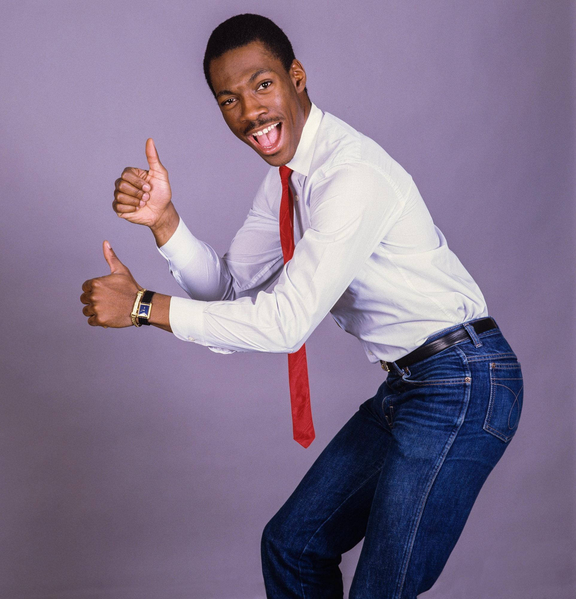 eddie murphy 桌布 壁紙 — 一個快樂的男人穿著白襯衫和紅領帶，在純紫色工作室背景前熱情地豎起兩個大拇指。