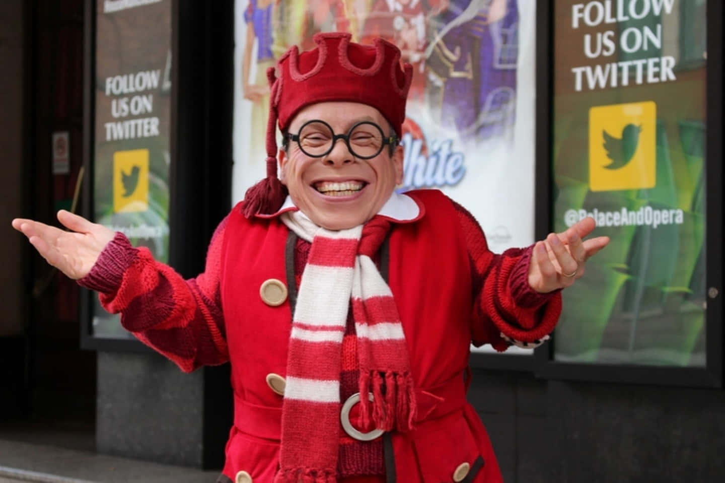 Warwick Davis 桌布 壁紙 — 一個快樂的男人穿著紅色皇冠外套和圍巾，在劇院外張開雙臂，笑容滿面。